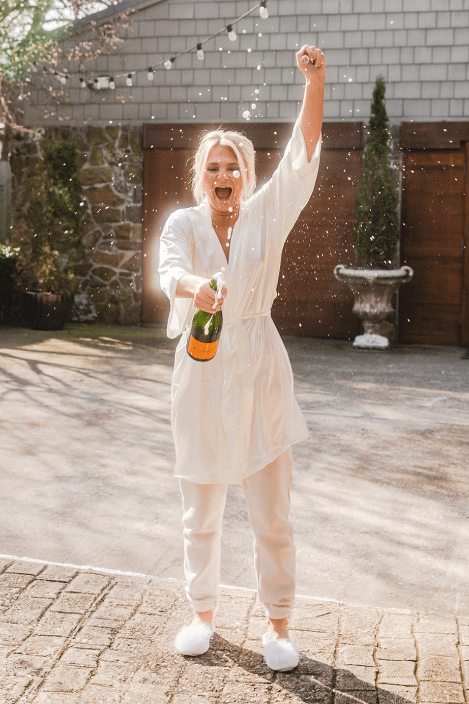 Woman in white pajamas celebrating outdoors with champagne, spraying foam, smiling, on a sunny day.