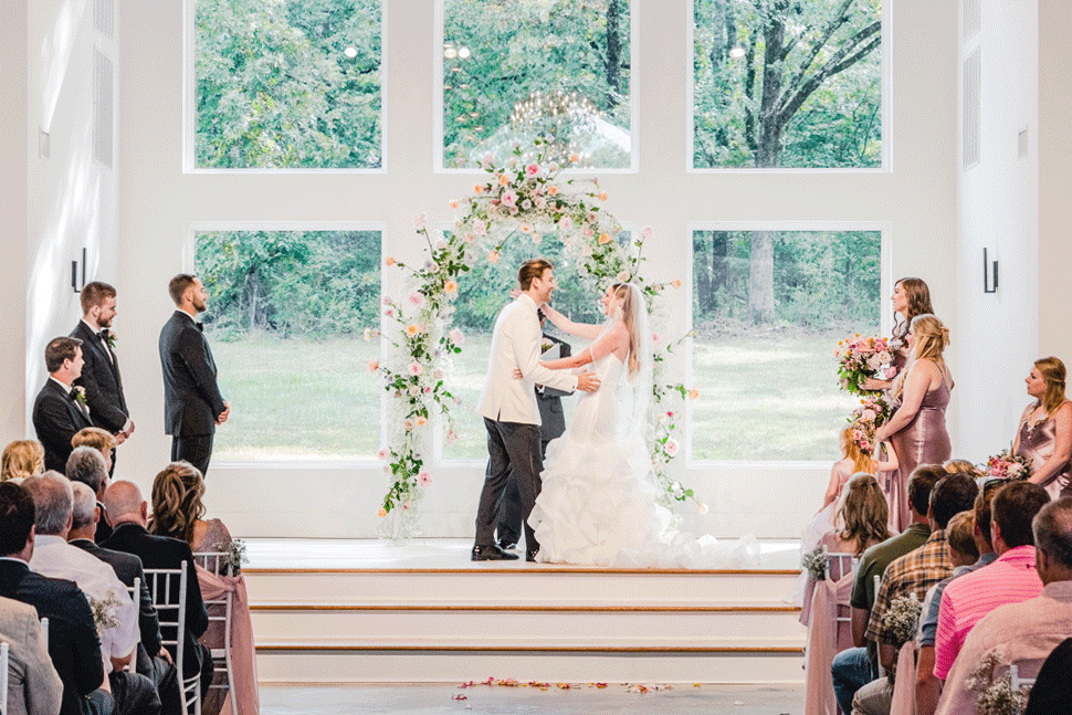 Bride and groom exchanging vows during wedding ceremony in a bright, modern church with large windows and family and friends watching.