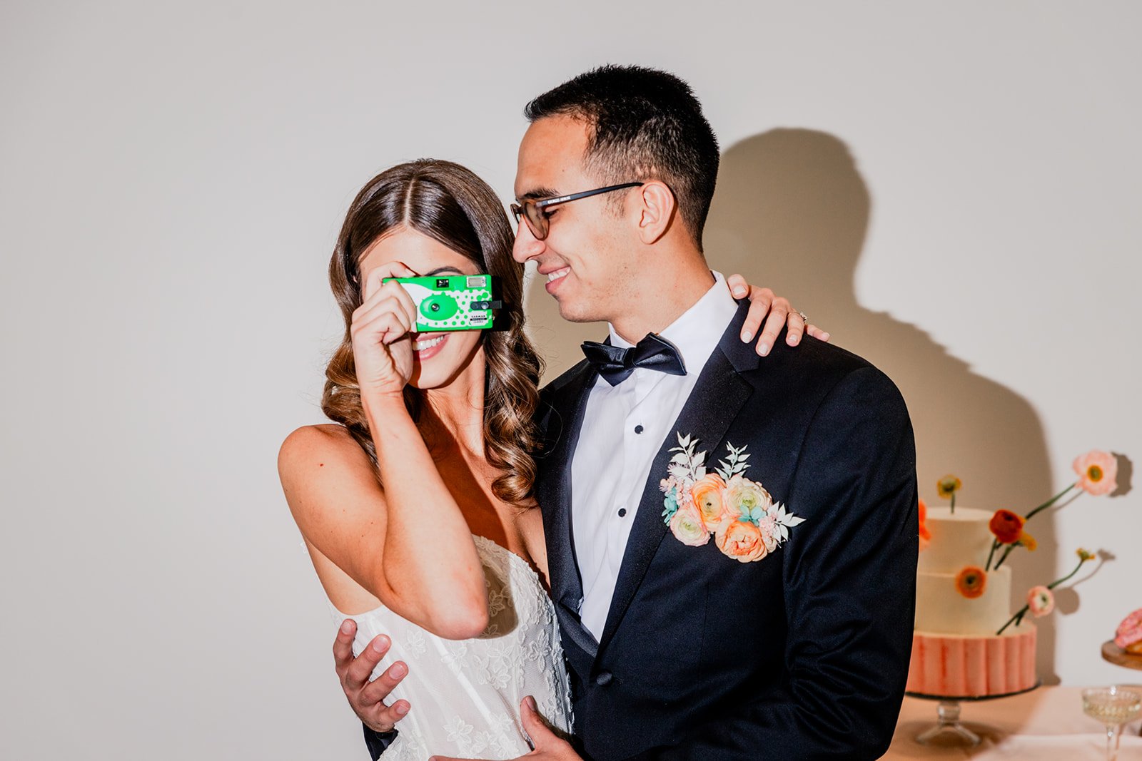 A bride and groom at their wedding, with the bride taking a photo with a green camera, both smiling.