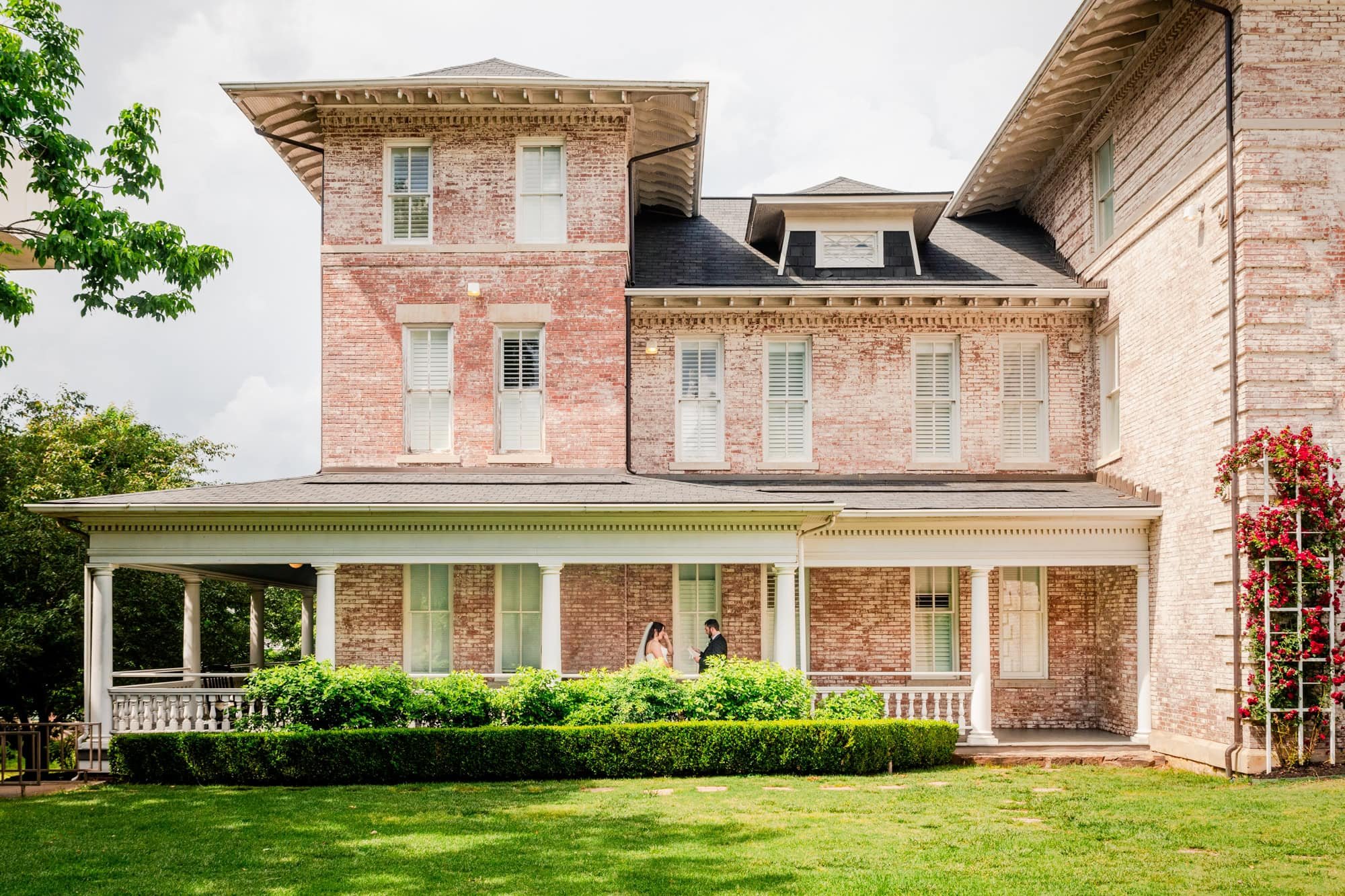 A couple getting married outside a large brick house with a front porch and lush green lawn.