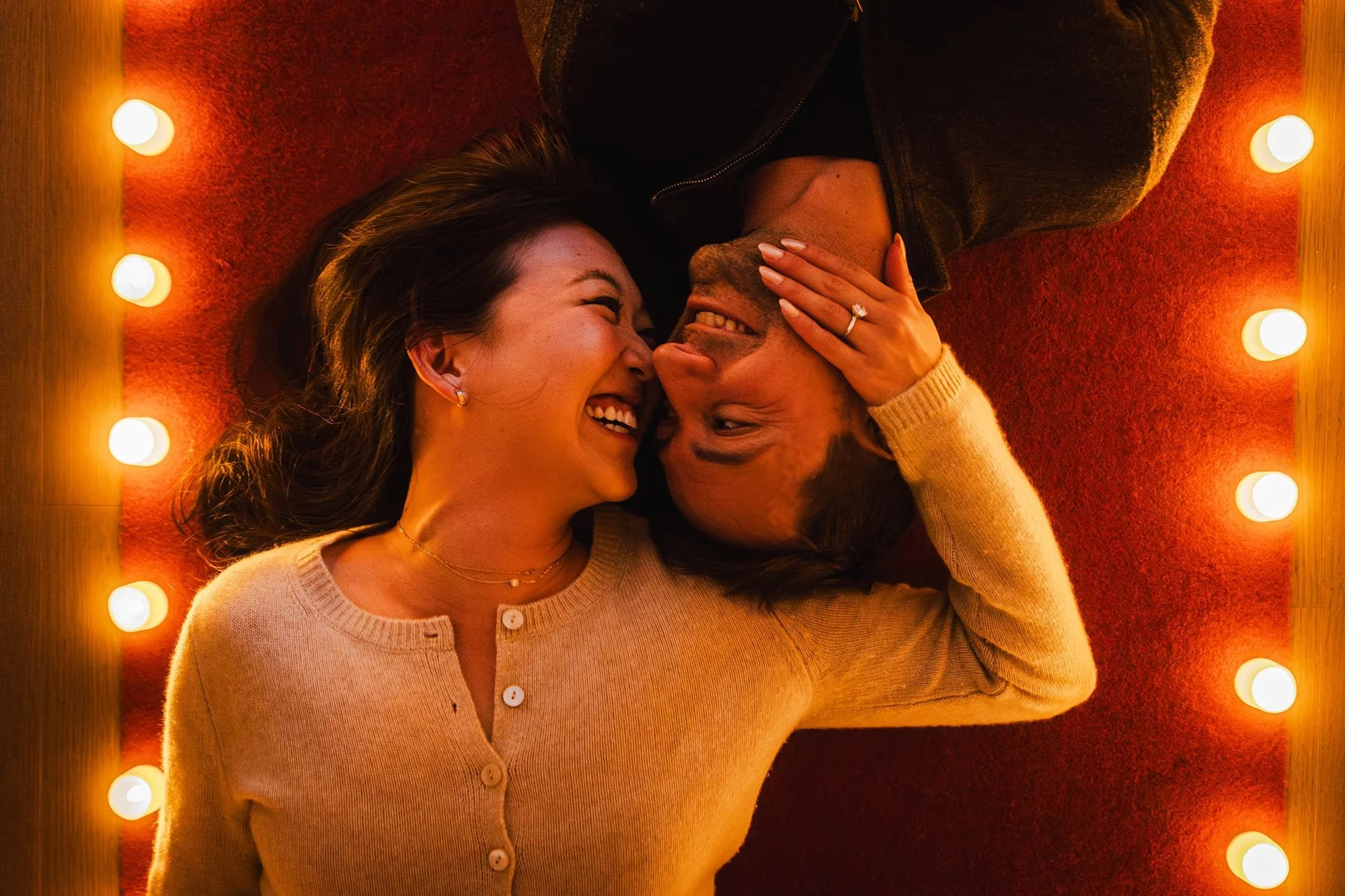 A couple lying on a red carpet surrounded by a string of light bulbs, smiling and looking at each other, with the woman touching the man's face.