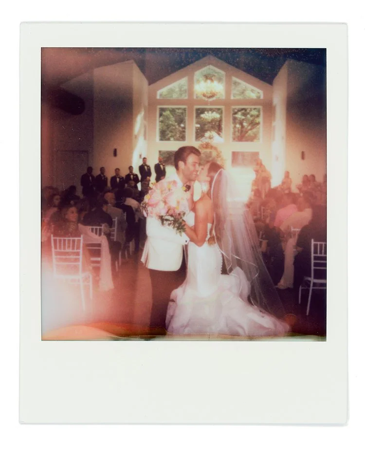 Photograph of a wedding ceremony with a bride and groom facing each other and holding hands in a church or hall, with guests seated in the background and large windows behind them.