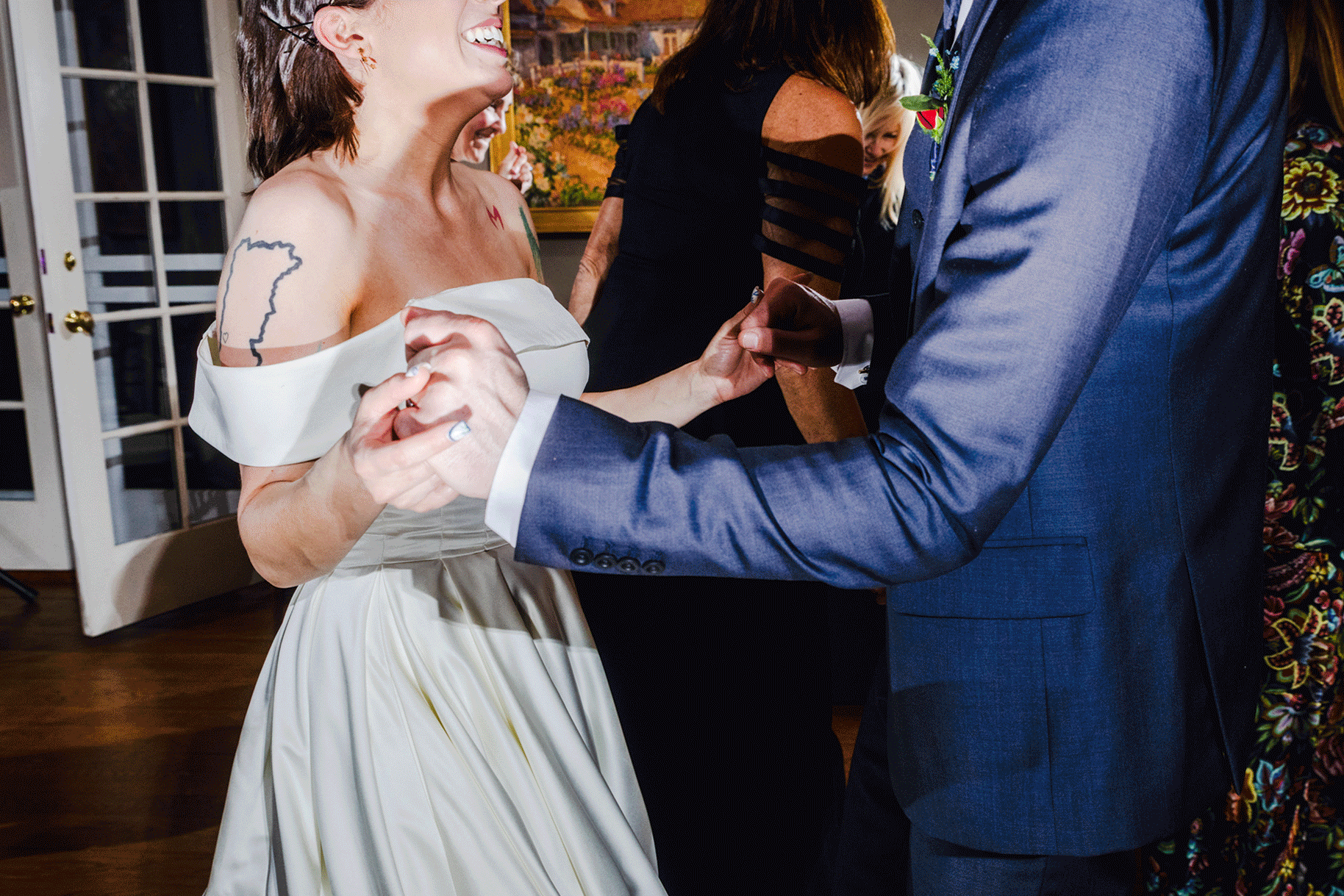 A woman in a white dress with tattoos on her arm is holding hands with a man in a blue suit during a wedding dance. They are smiling and looking at each other, with other people in the background at a wedding reception.