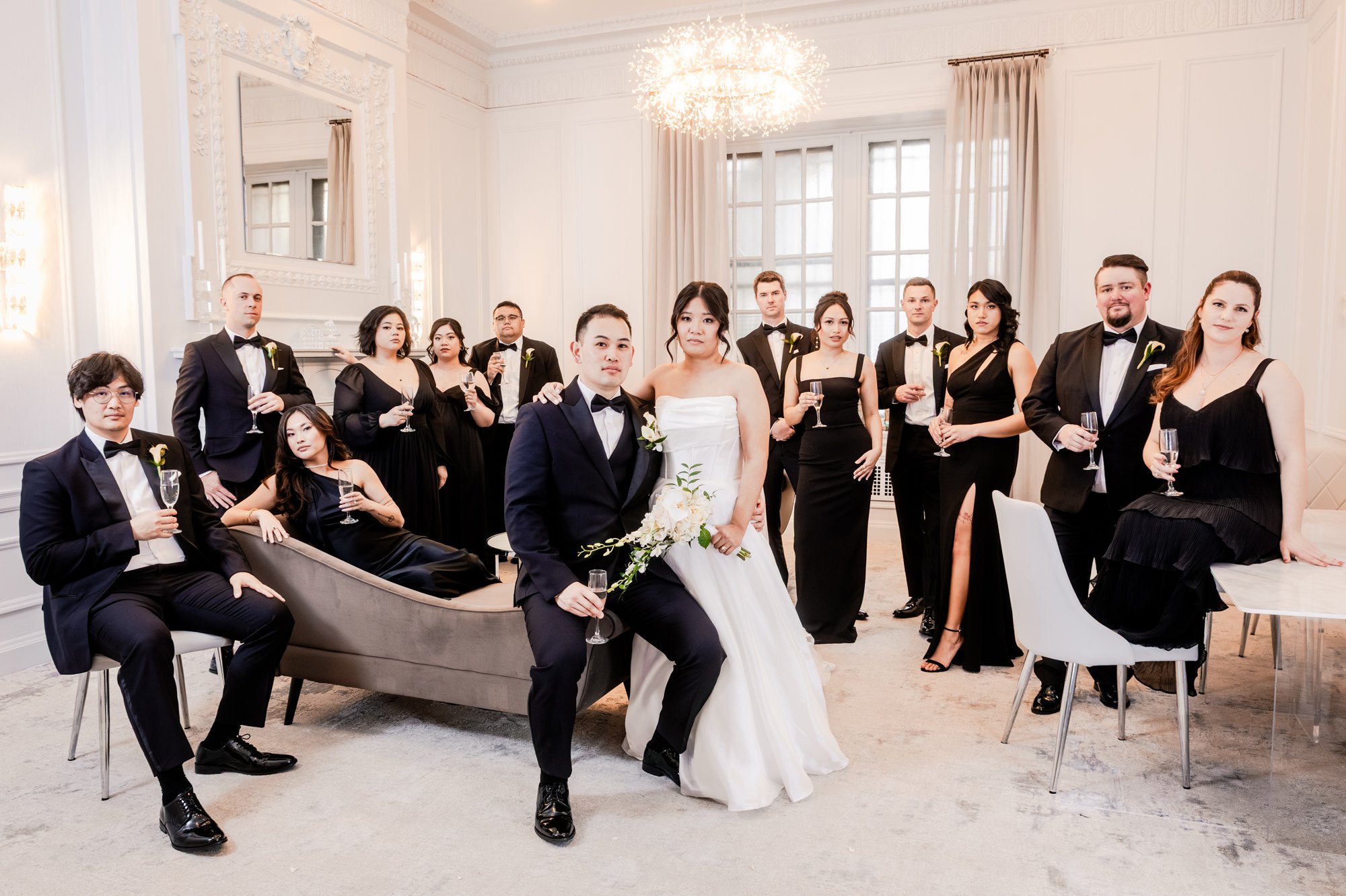 A wedding party with the bride and groom seated in the center, surrounded by their bridesmaids and groomsmen dressed in formal black attire, inside an elegant, well-lit room with white walls, large windows, and modern decor.