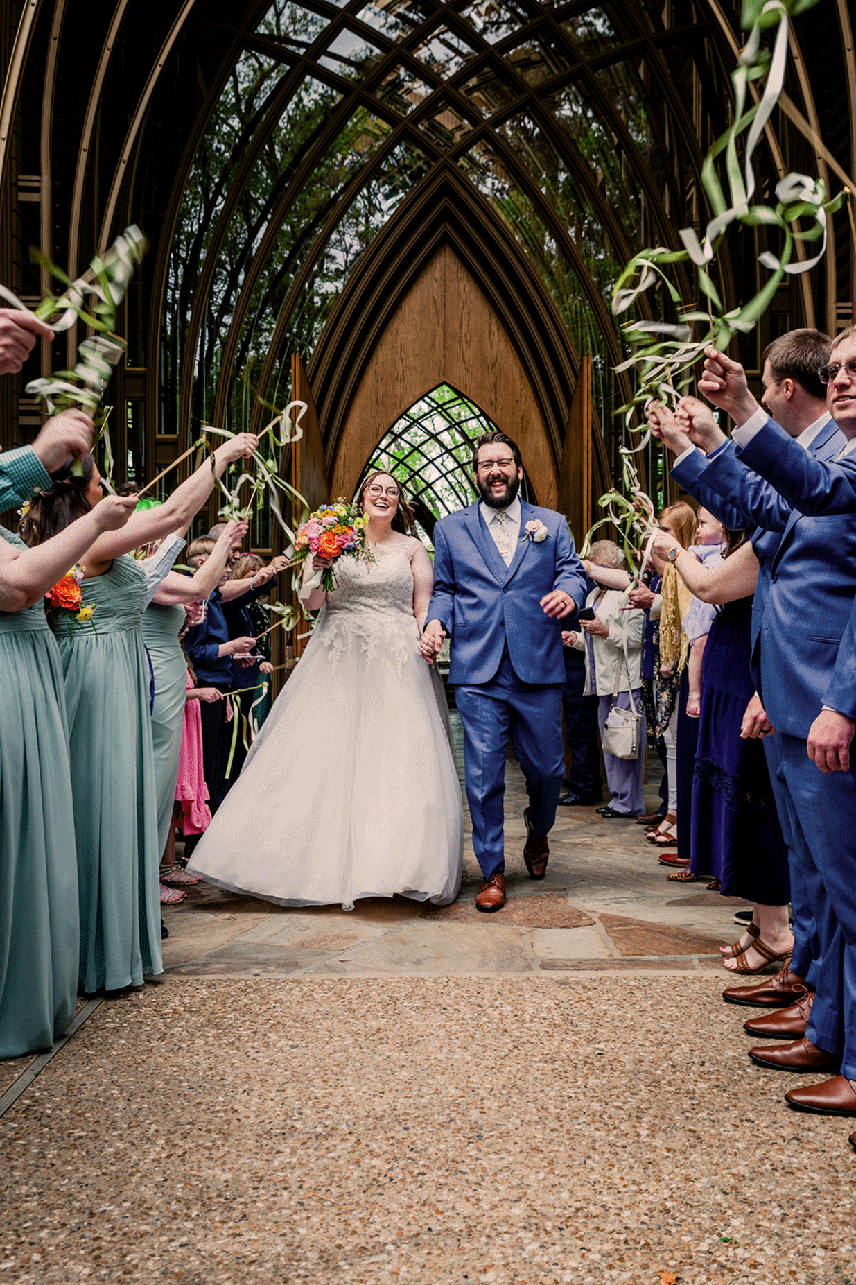 Bride and groom walking down the aisle after wedding ceremony, surrounded by guests holding streamers in an indoor venue with wooden architectural design.