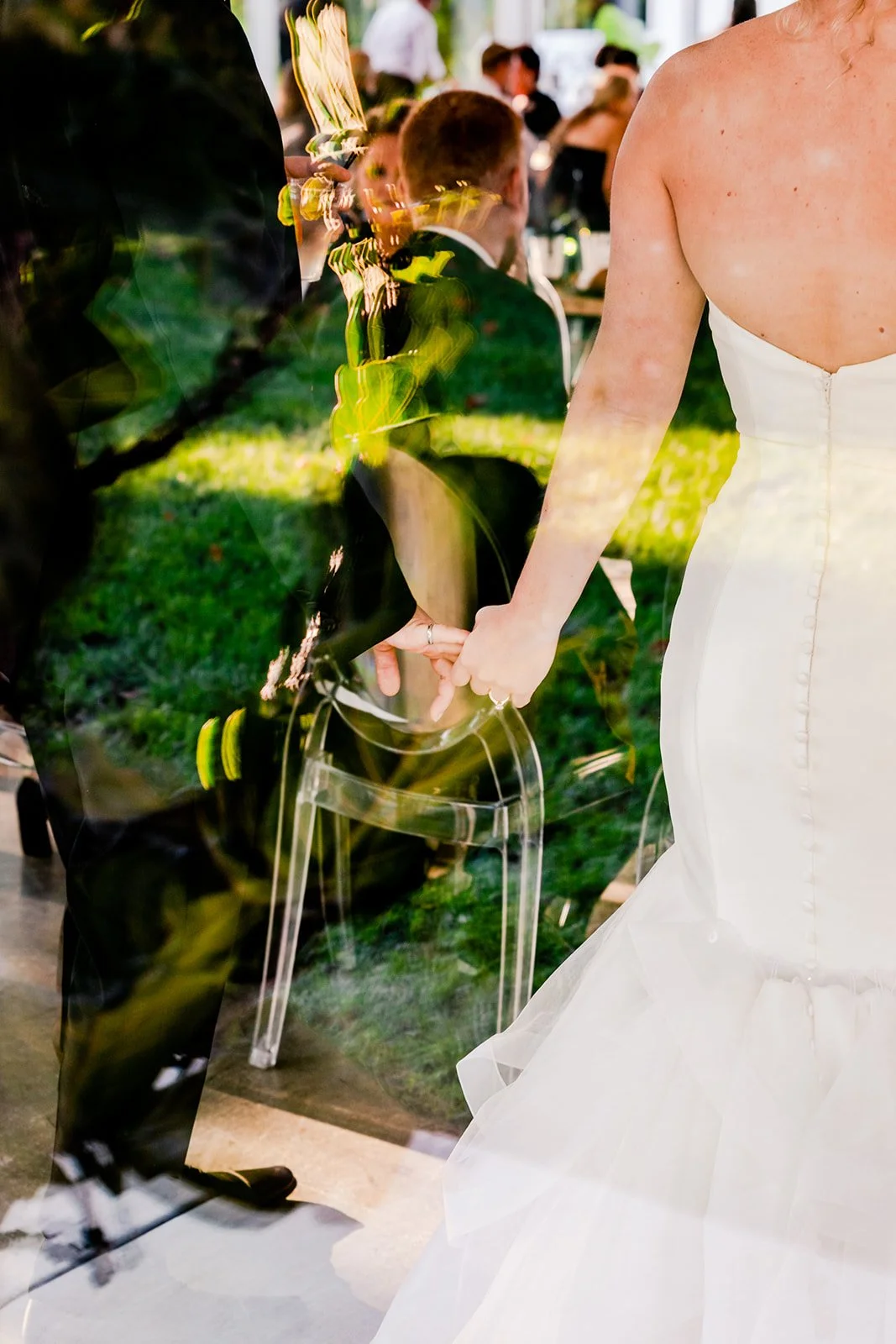 A couple holding hands at a wedding ceremony, with the bride wearing a white strapless gown and the groom in a black suit, reflected in a glass window.