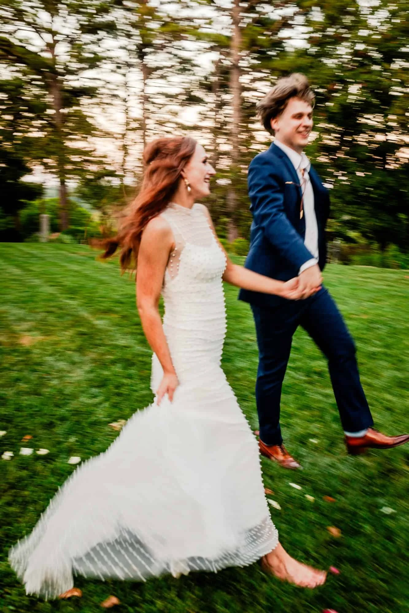 A happy couple in wedding attire walking hand in hand outdoors on grass. The bride is in a white gown, and the groom is in a navy blue suit. The background shows trees and a sunset sky.
