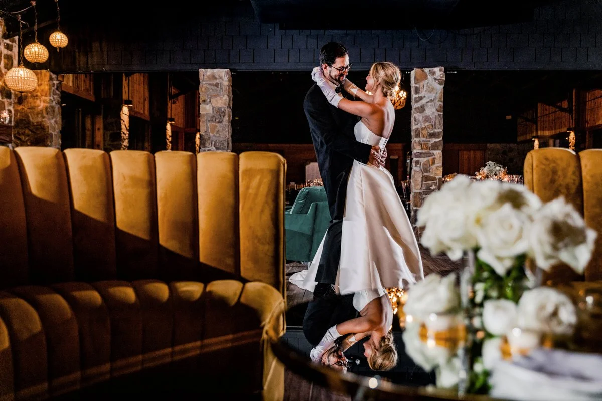 A bride and groom dancing together in a warmly lit, rustic wedding venue with stone columns, wooden accents, and elegant decor.