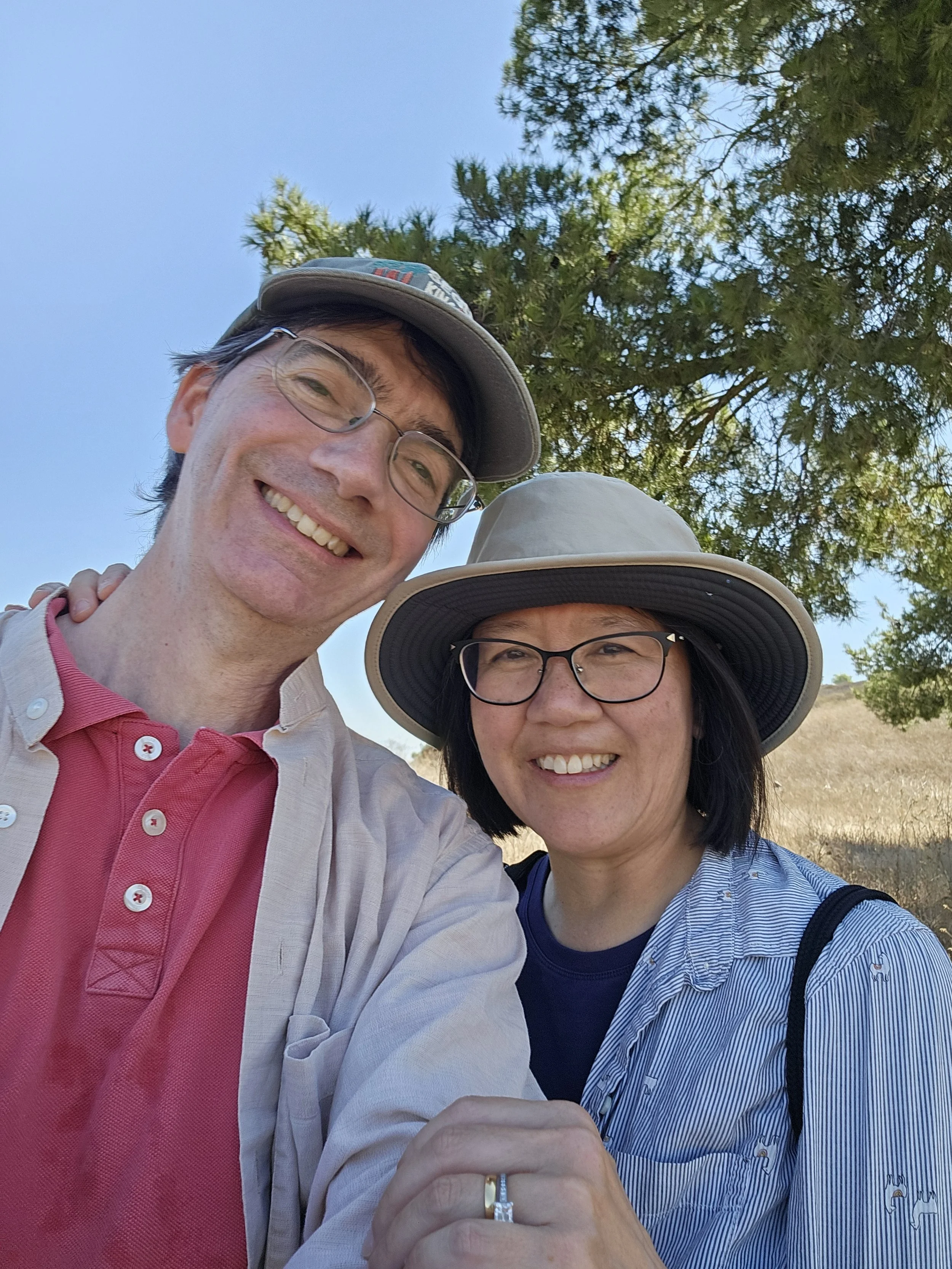 A smiling woman wearing glasses and a blue shirt embraces a man smiling in a red shirt and a ball cap while standing in the shade of a tree.