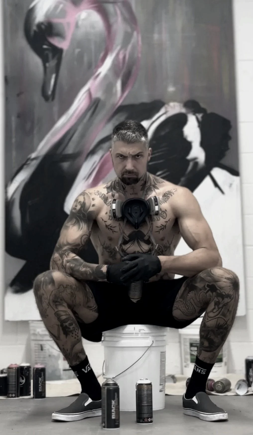 A muscular man with tattoos is squatting with a serious expression in front of a large African painted art piece, holding a spray paint can, with various cans on the floor.