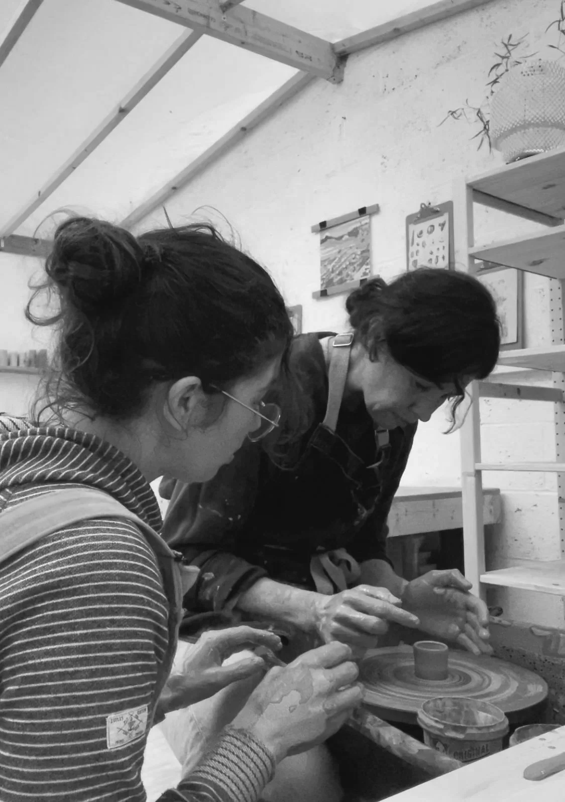 Two women working together on a pottery wheel in a studio, shaping clay with their hands.