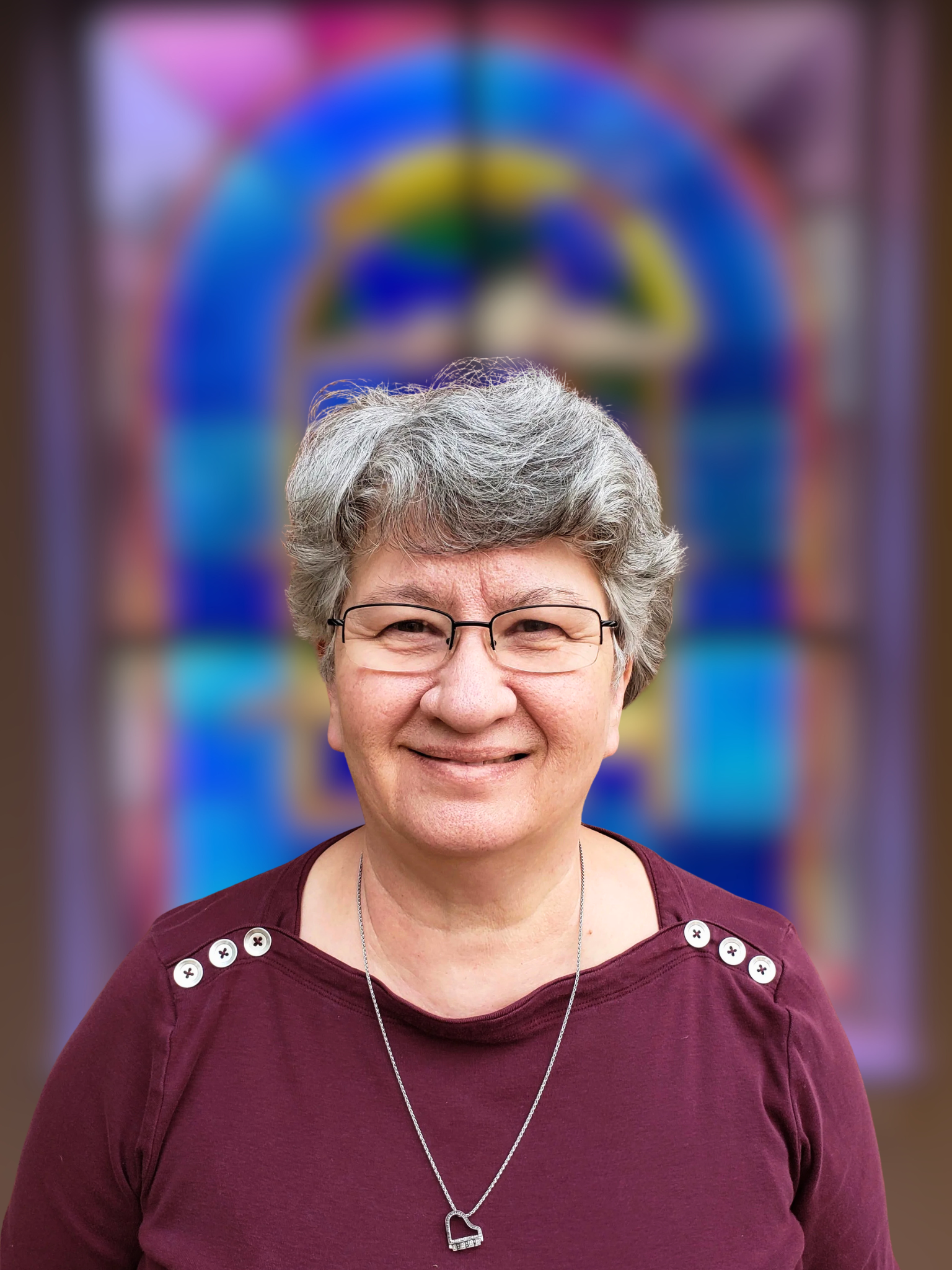 Close-up of a smiling woman with short gray hair, glasses, wearing a maroon top with buttons on the shoulders, standing in front of a brick wall.