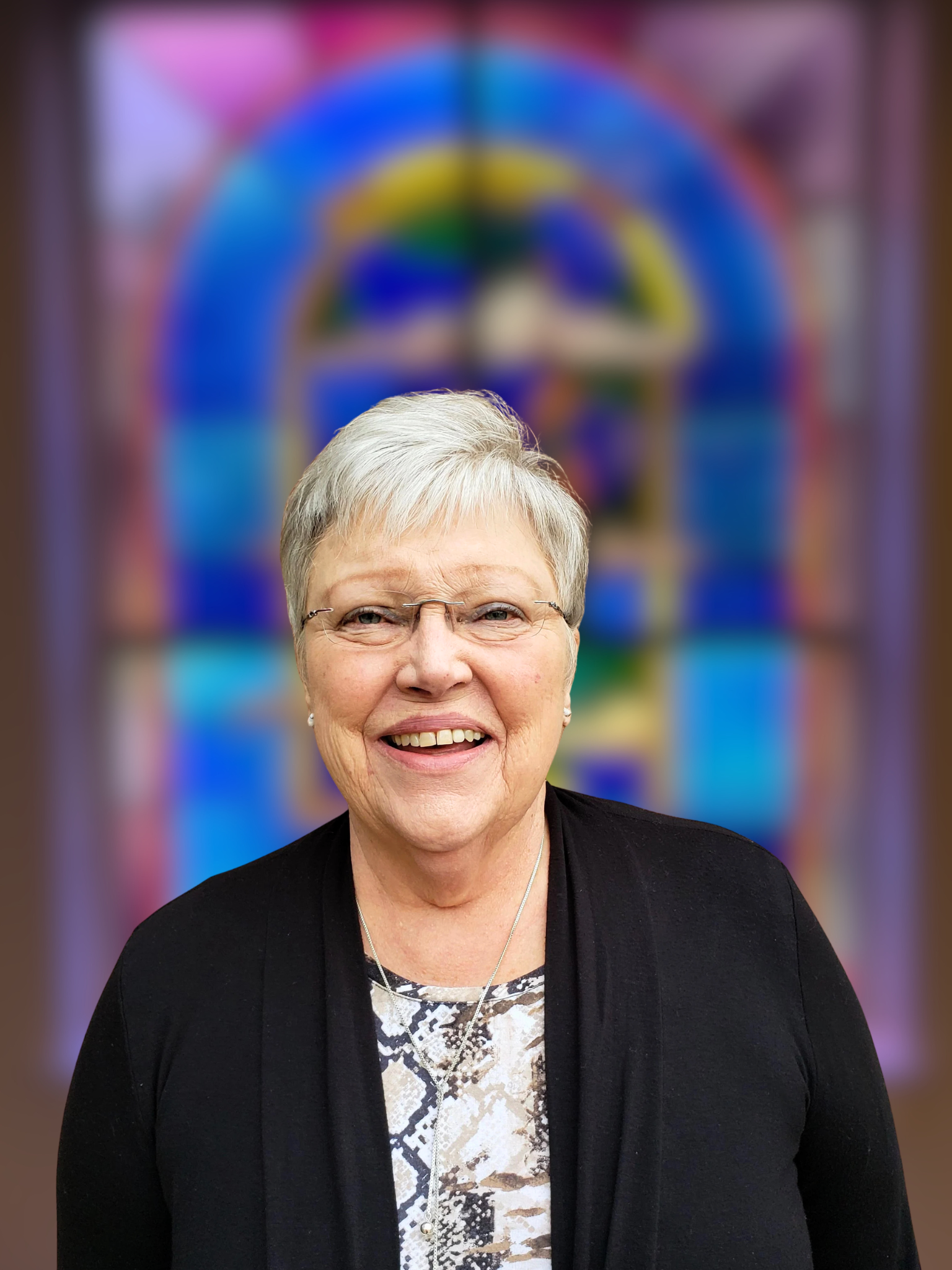 Close-up of a smiling elderly woman with short white hair and glasses, wearing a black blazer and patterned top, standing in front of a brick wall.