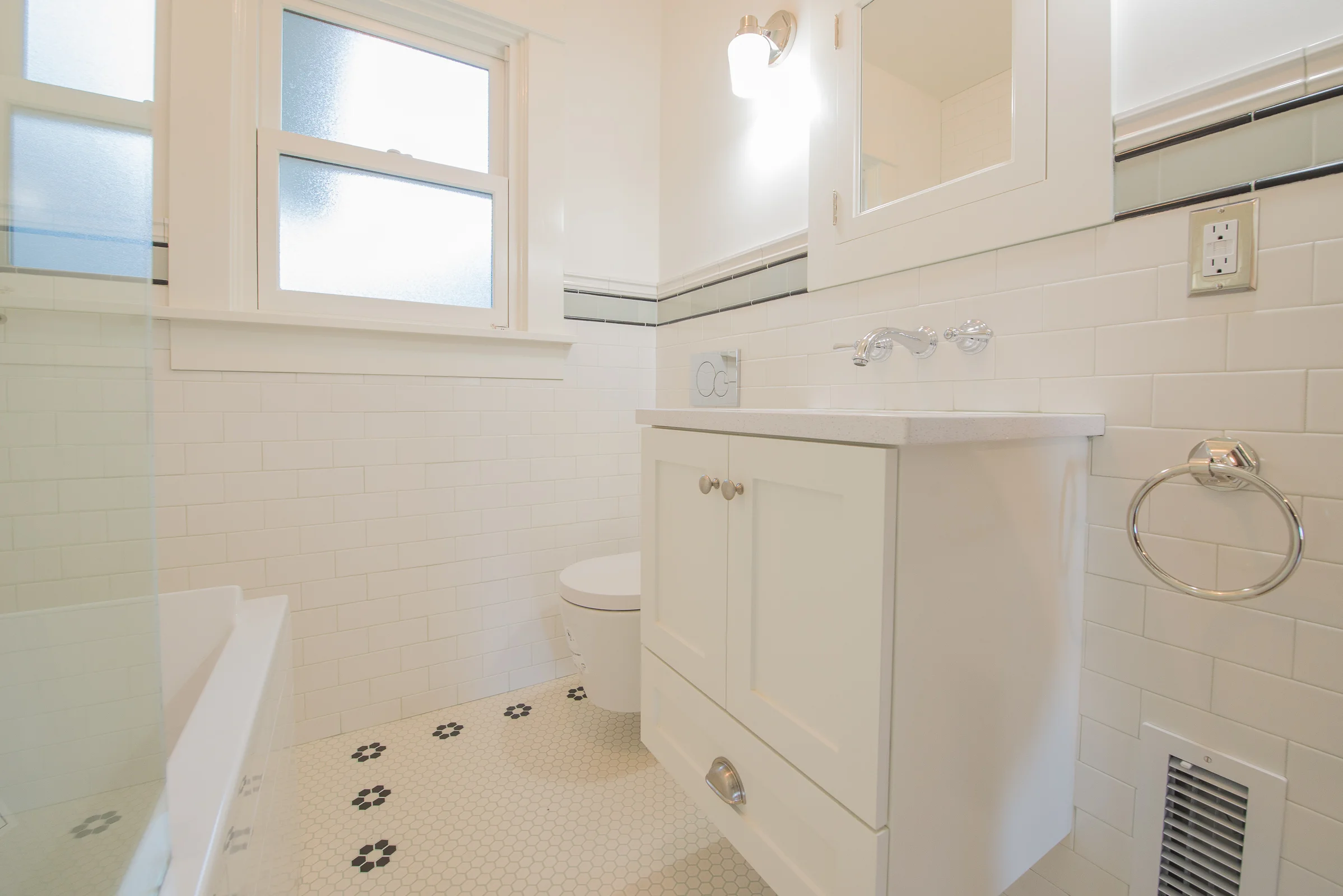 A neutral yet detail-focused package was requested by the clients for the finishes. Our goal was to make a statement in this tiny bathroom without the overwhelming use of a bold color palette. The contrast of multiple white textures—hex floors, wall tile, cabinetry, deep soaking tub and countertops—lend a sense of interest. The bathroom utilizes the most subtle of space planning techniques: a wall hung toilet and faucet save several inches of room. The floating vanity, extra narrow and extra-deep soaking tub—paired with a glass partition instead of a shower curtain—all add to the perception that there is an ample amount of floor space.