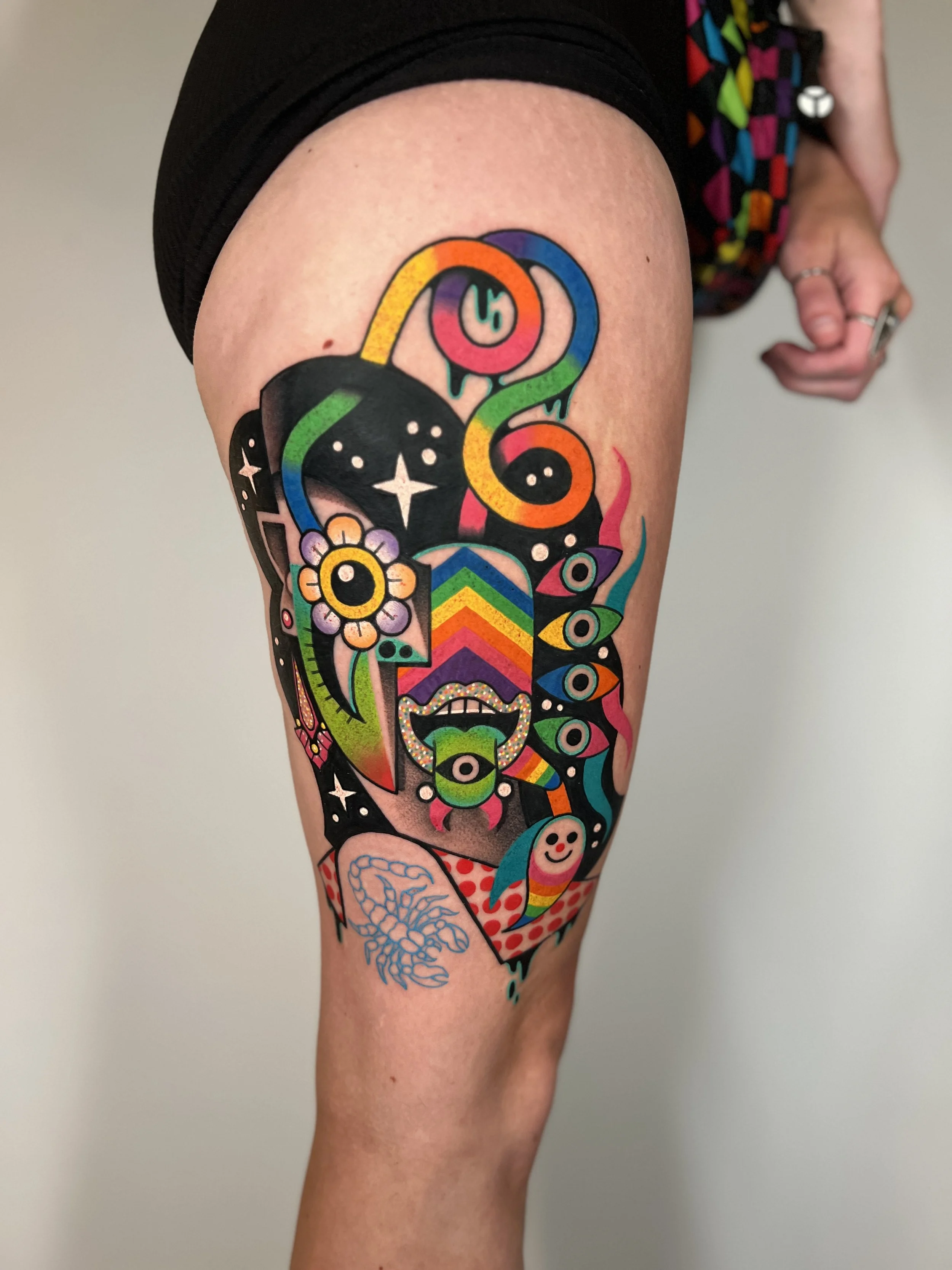 Colorful tattoo on person's thigh, featuring abstract and surreal elements including multiple eye shapes, a flower, a smiling face, and swirling patterns in bright colors.