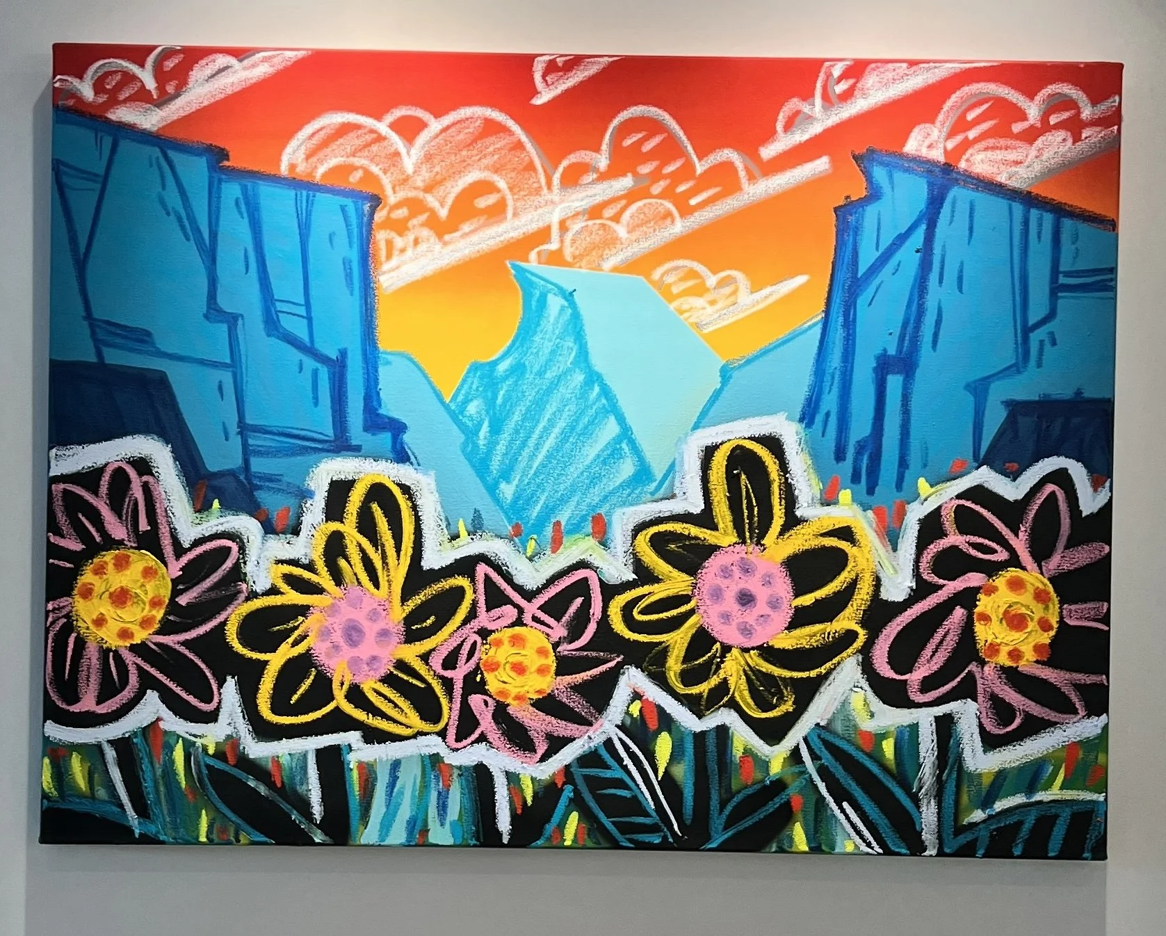 Colorful abstract painting showcasing a mountain range in shades of blue with a vibrant sky in orange and yellow, surrounded by bright flowers with pink, yellow, and purple centers, and black outlines with green leaves at the bottom.