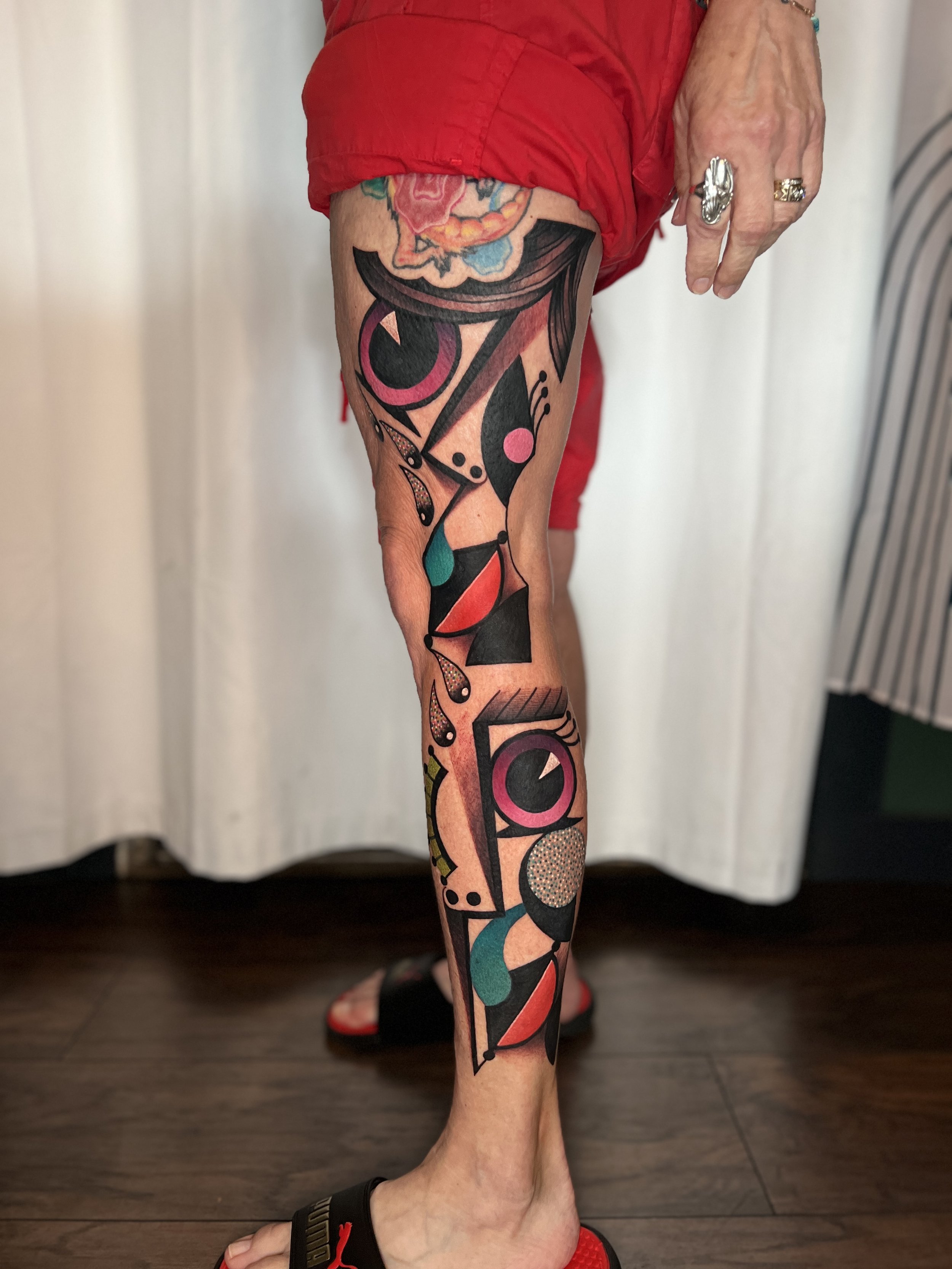 A person's lower leg with a large, colorful abstract tattoo featuring geometric shapes, bold lines, and bright colors including pink, red, black, teal, and yellow. They are wearing red shorts, black slides, and have multiple rings on their fingers.