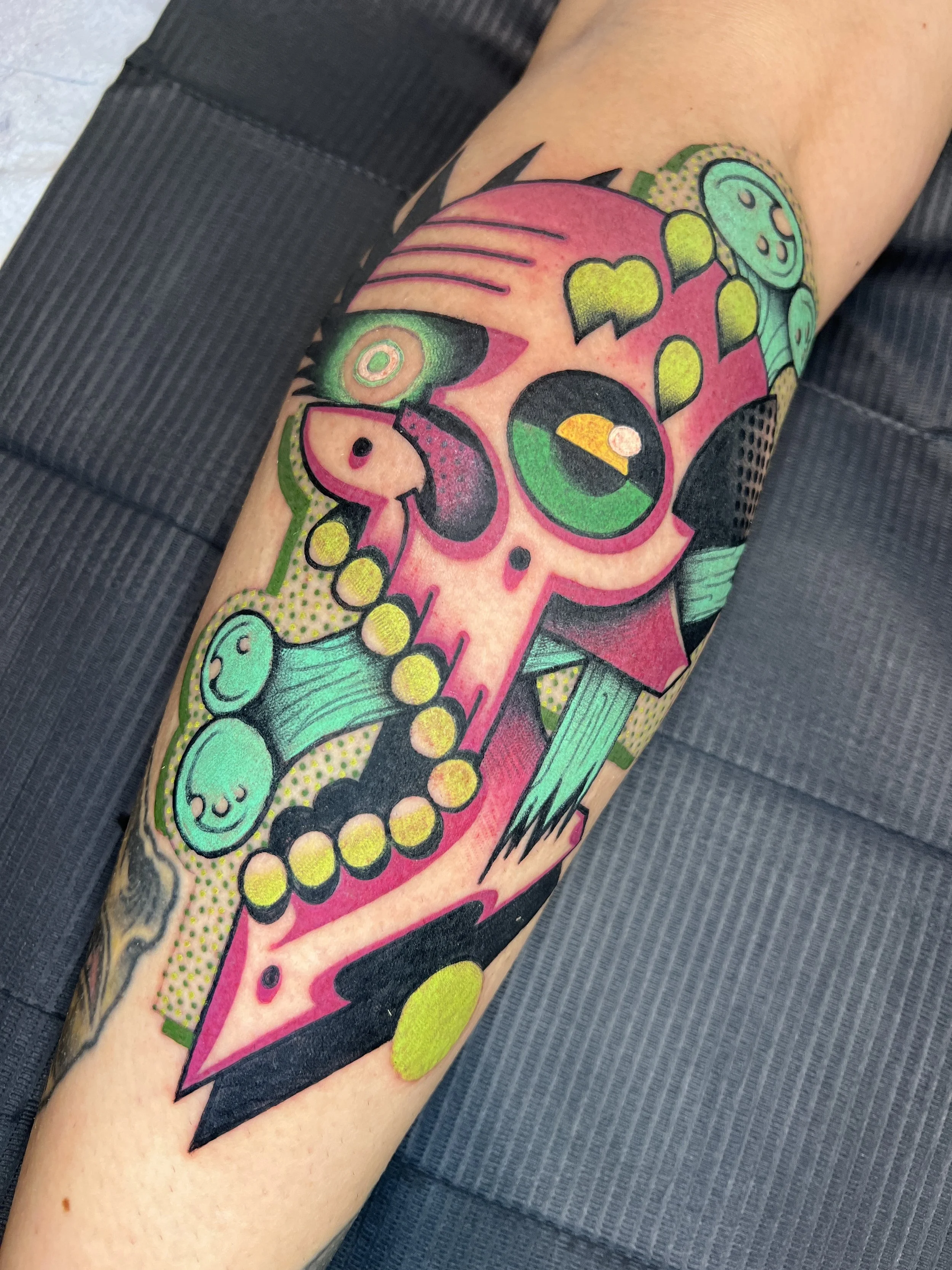 Colorful tattoo of a pink skull with green and yellow accents, surrounded by abstract shapes and patterns in green, teal, and yellow.