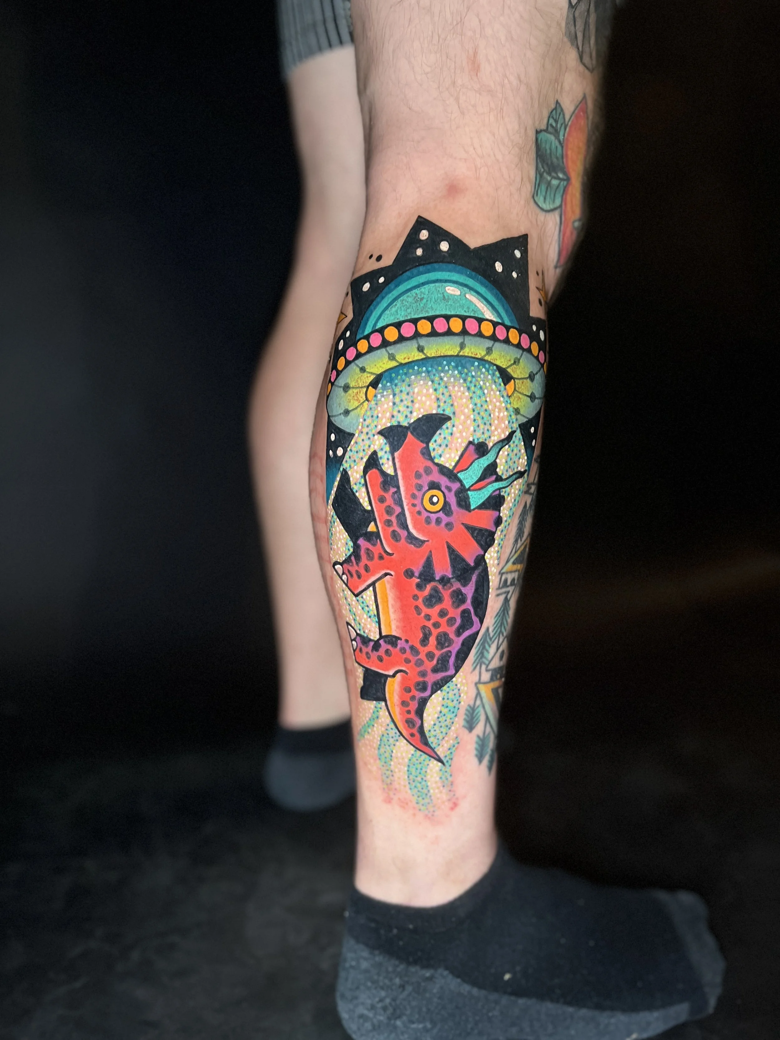 Colorful tattoo of a red and purple dinosaur preparing to eat a flying fish with a halo above, set against a background of a glowing, stylized UFO.