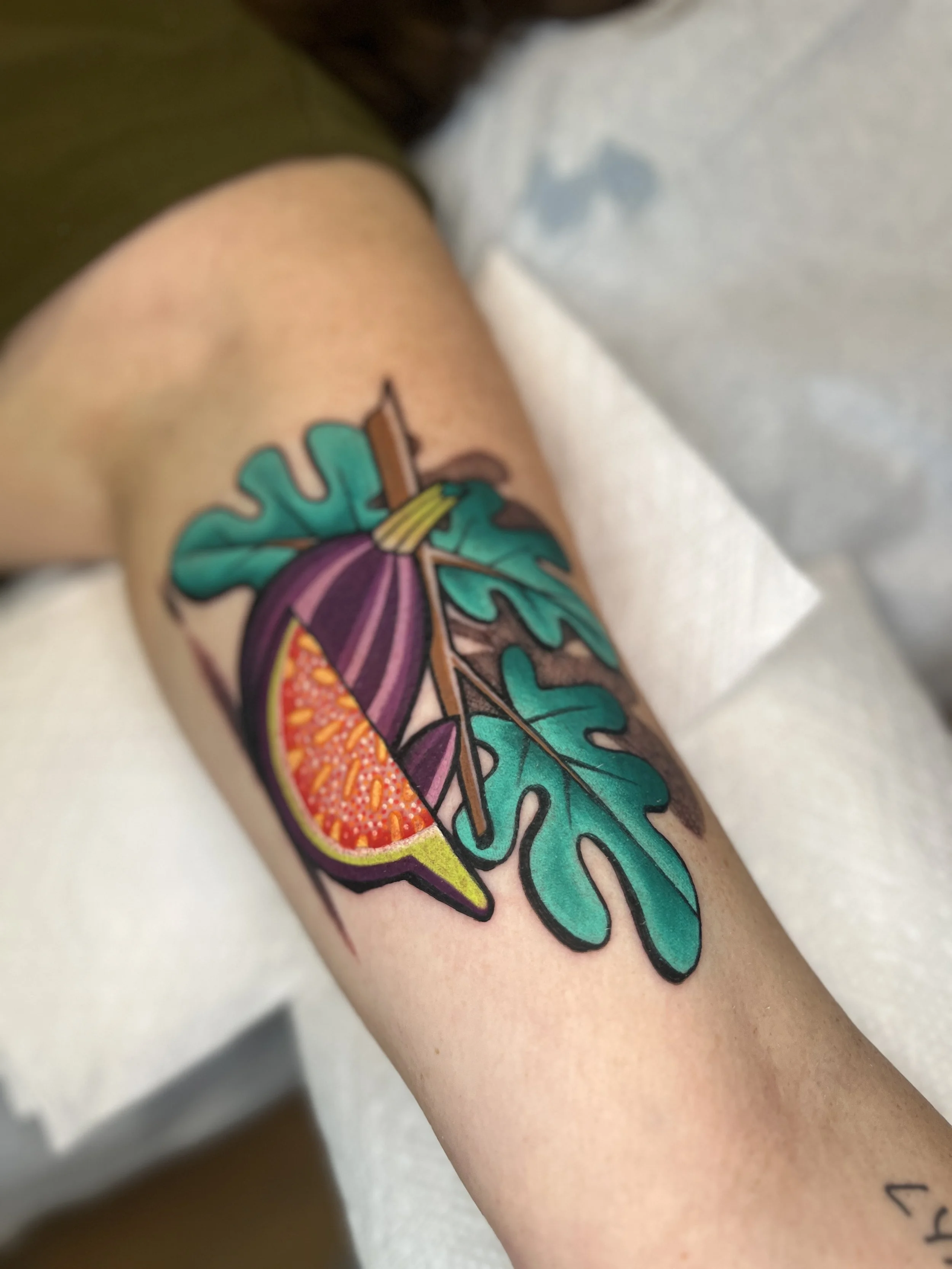 Colorful tattoo on a person's arm featuring a fig cut in half with interior details, large green leaves, and a brown stem in a bold, illustrative style.