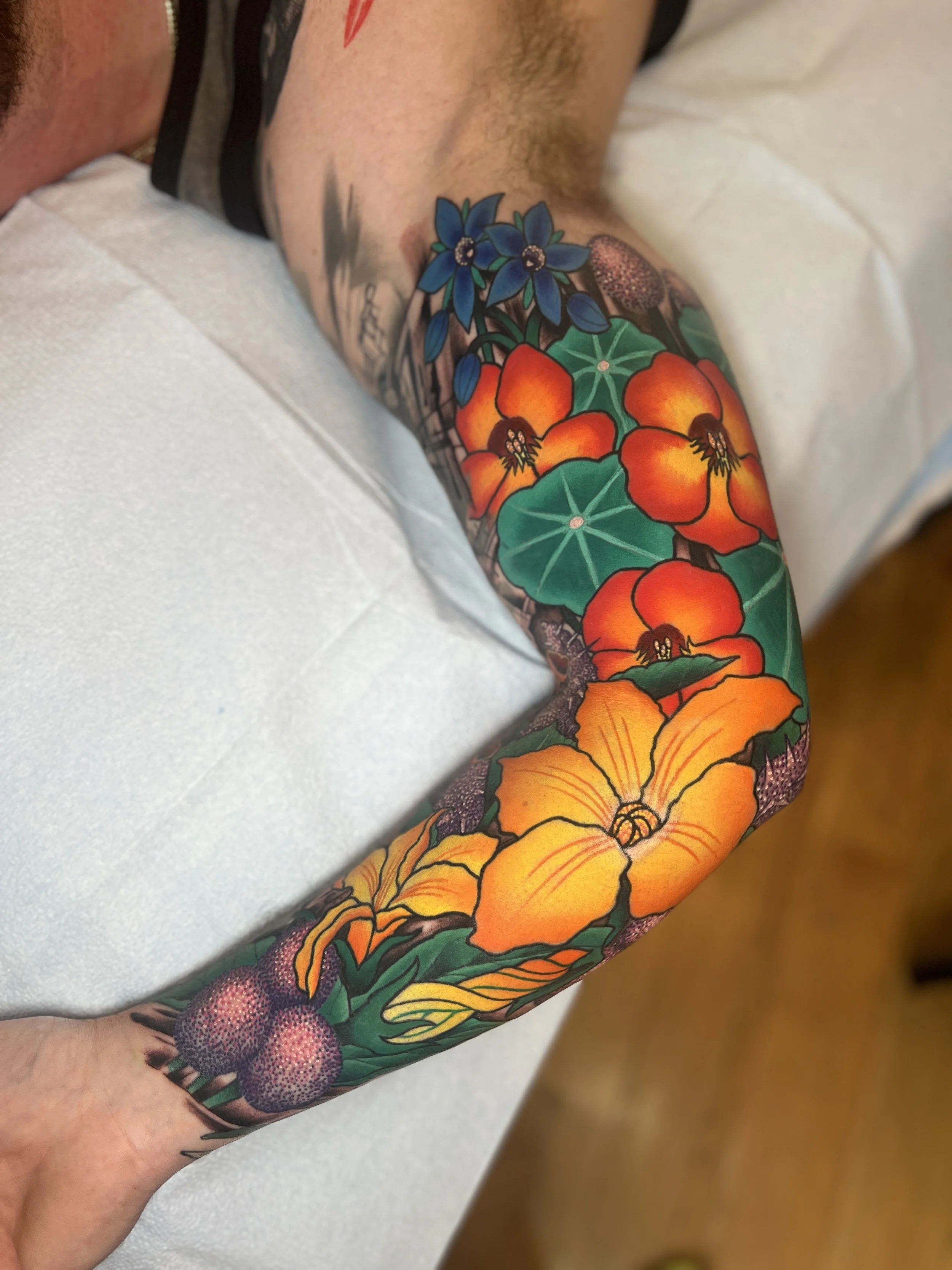 Colorful tattoo of various flowers, including orange and yellow blooms, with green leaves, on a person's arm.