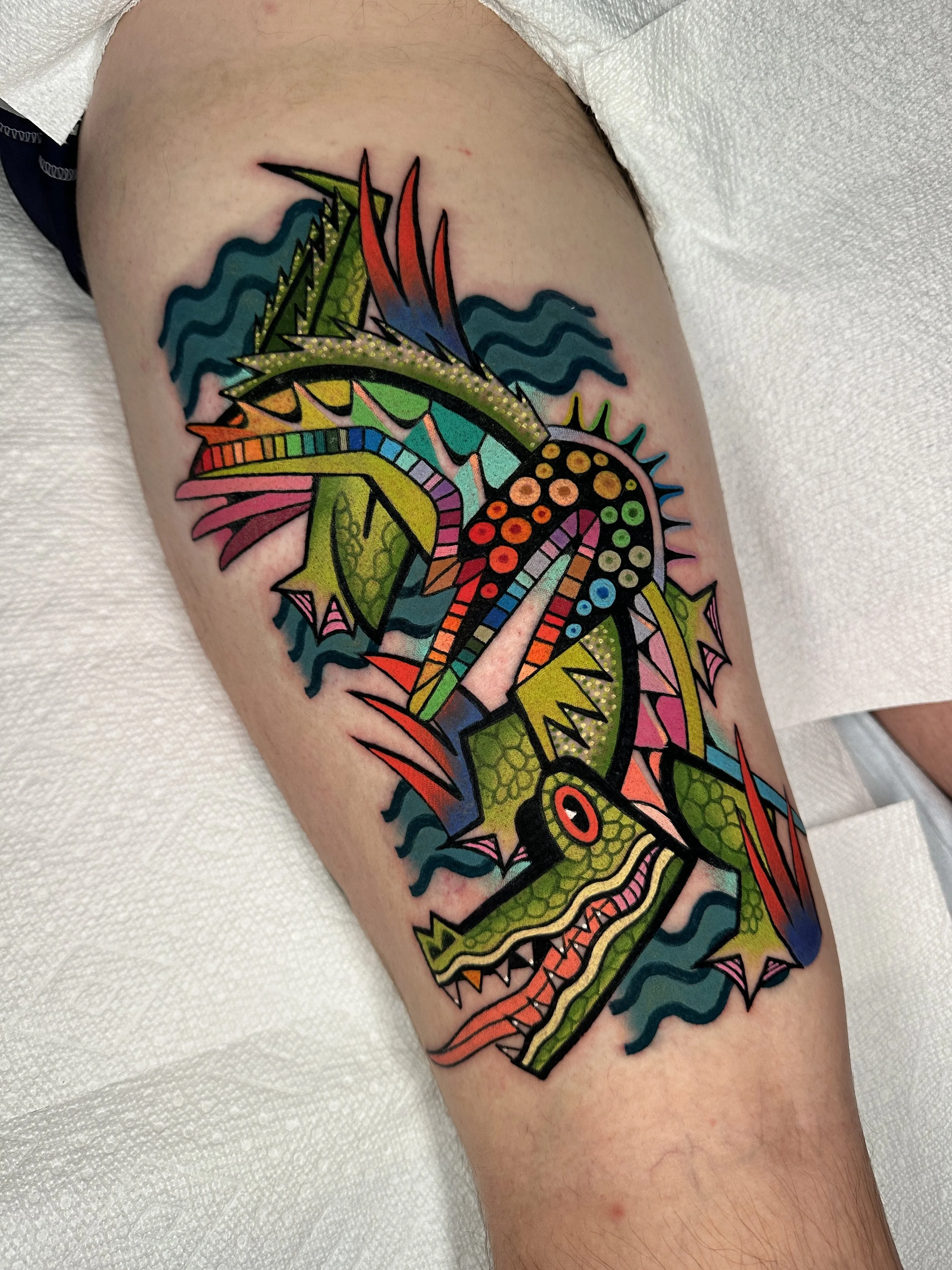Colorful tattoo of a dragon with sharp, jagged features, vibrant patterns, and bright colors including green, red, yellow, pink, and blue on the person's thigh.