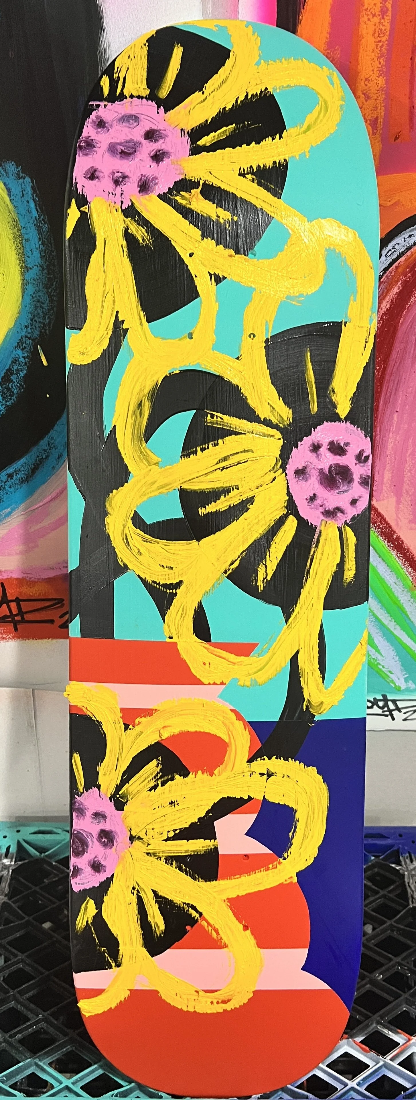 Colorful skateboard deck with abstract art featuring bright yellow, pink, black, turquoise, red, white, and blue brushstrokes, with pink flower-like shapes.