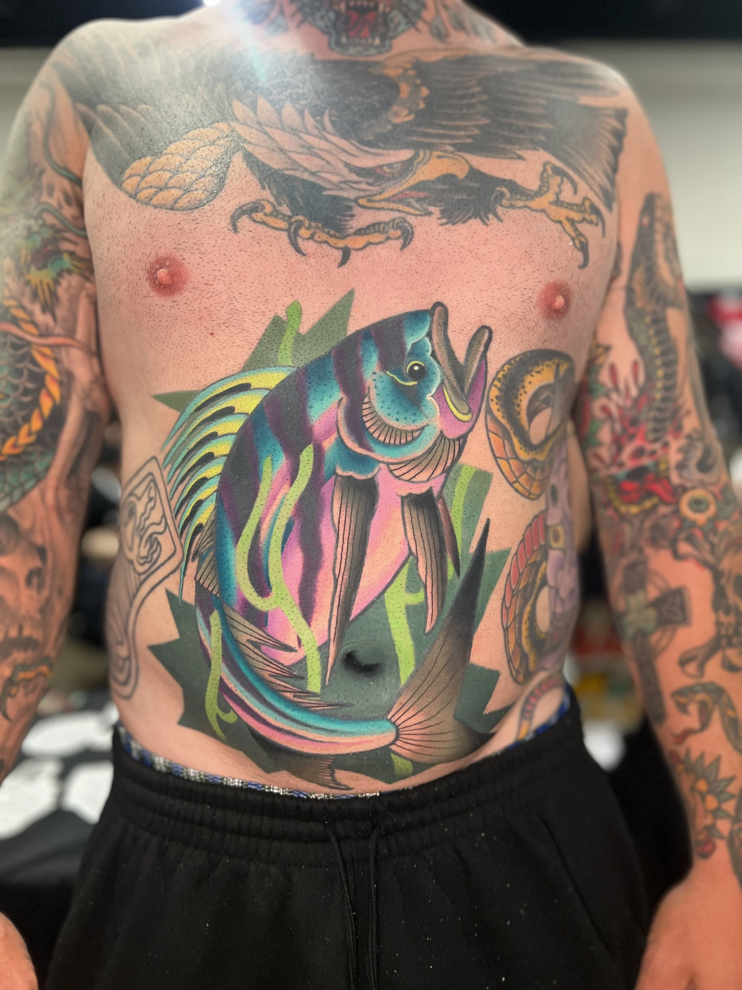 Colorful tattoo of a fish on a man's chest with other traditional-style tattoos on his arms and shoulders.