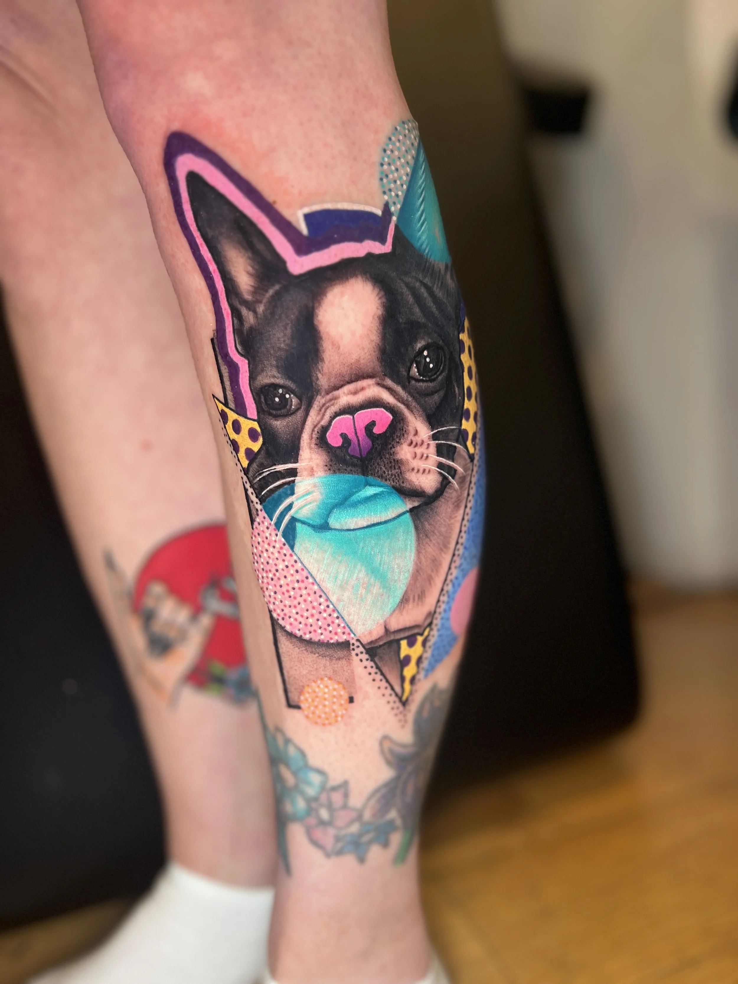 Colorful tattoo of a French Bulldog puppy's face, featuring vibrant geometric shapes and patterns in pink, blue, yellow, and purple on the person's leg.