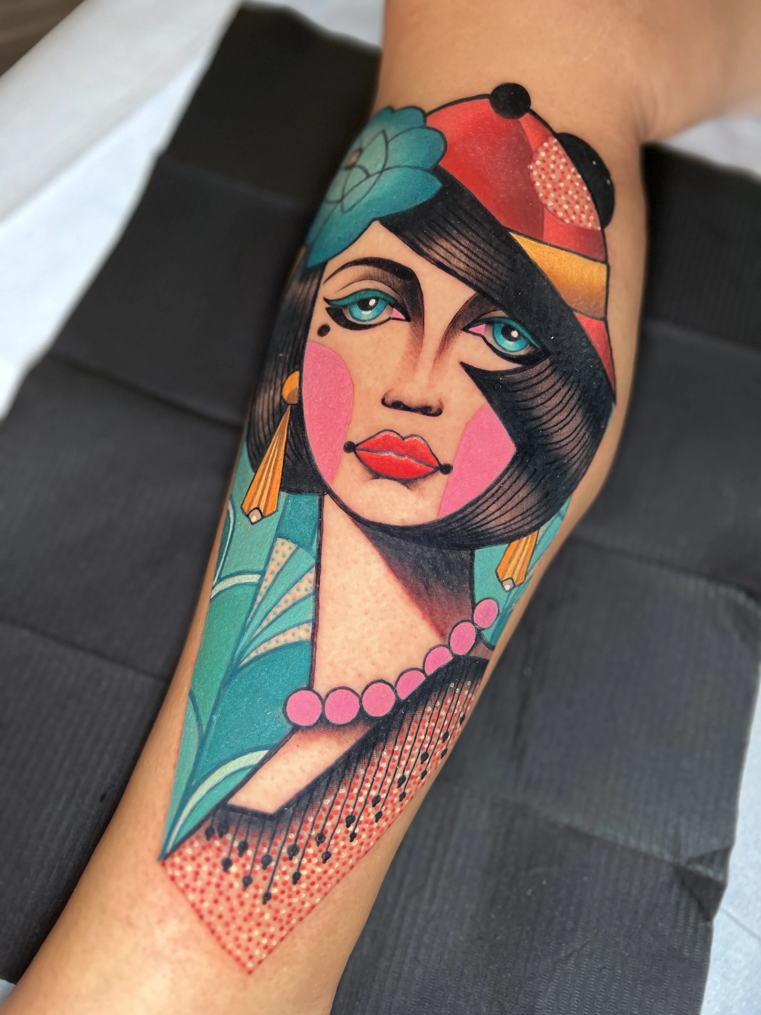 Colorful tattoo of a woman with teal eyes, black hair, pink cheeks, and red lips, wearing a red and orange hat, a pink pearl necklace, and earrings, with an abstract background.