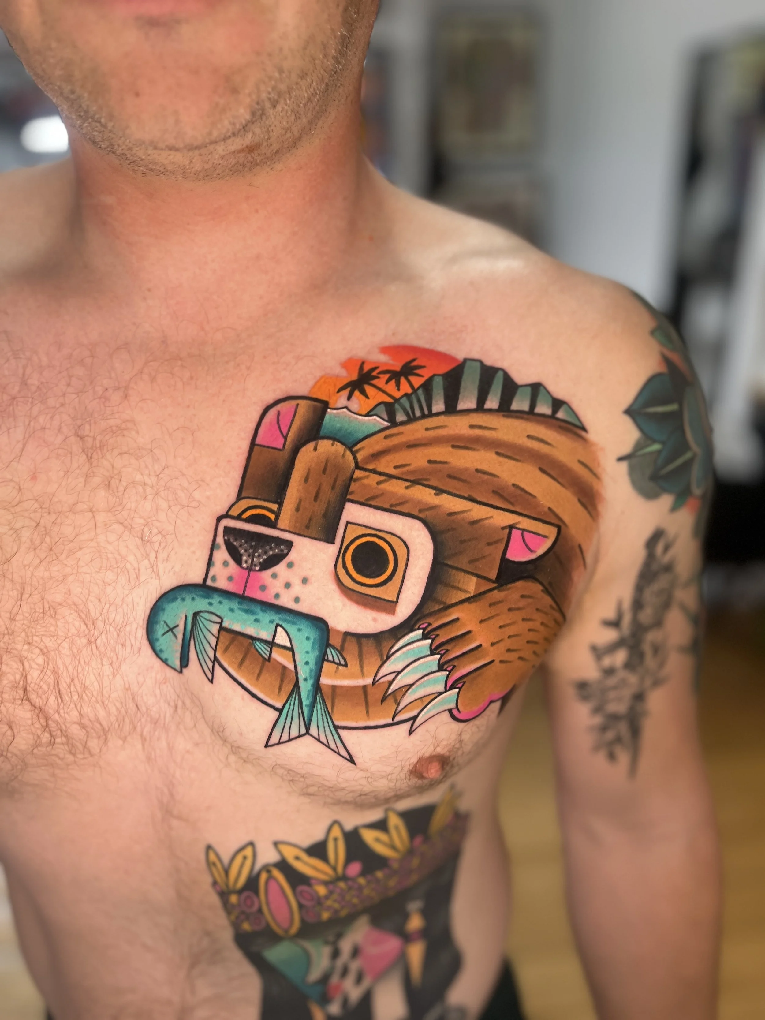 Colorful tattoo of a bear with a fish in its mouth on a man's chest, with a sunset and palm trees in the background.