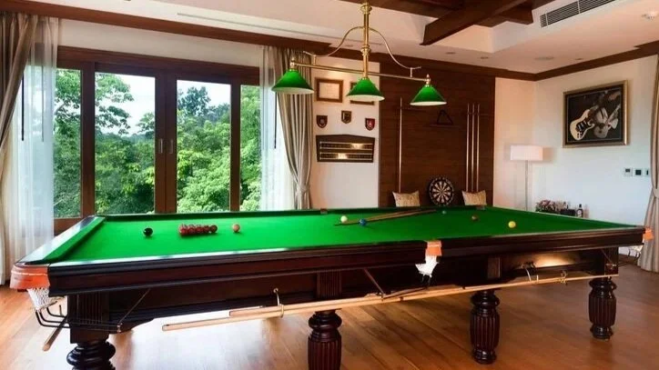 A luxurious game room with a green felt billiards table, hanging green pendant lights, a dartboard, artwork, and large windows showing a green outdoor landscape.