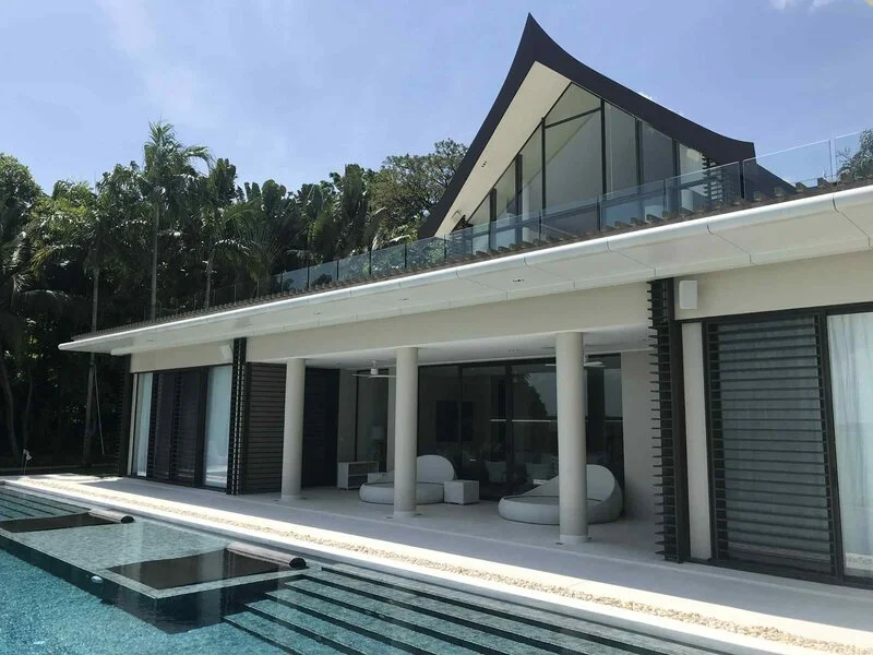 Modern two-story house with large glass windows, a balcony with glass railing, and a swimming pool in front, surrounded by lush greenery and tall trees.