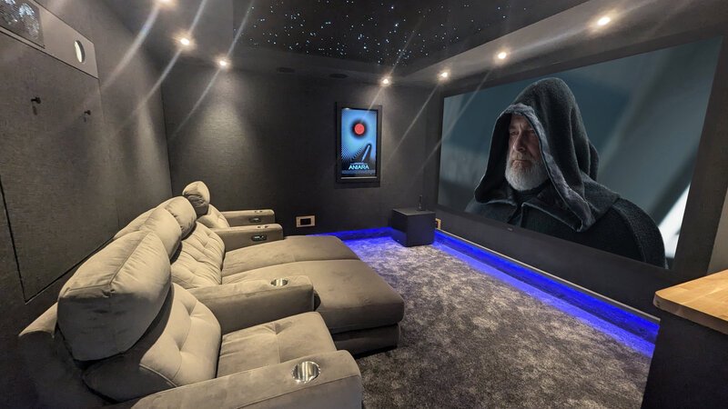 A modern home theater room with plush recliner seating, a large screen displaying a scene from a movie featuring an older man with a hoodie, dark accent walls, and starry ceiling lighting.