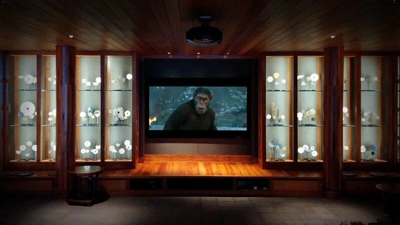 A home theater room with a large screen showing a monkey from a movie. The room has wooden walls, shelves with decorative objects, and a dark floor.