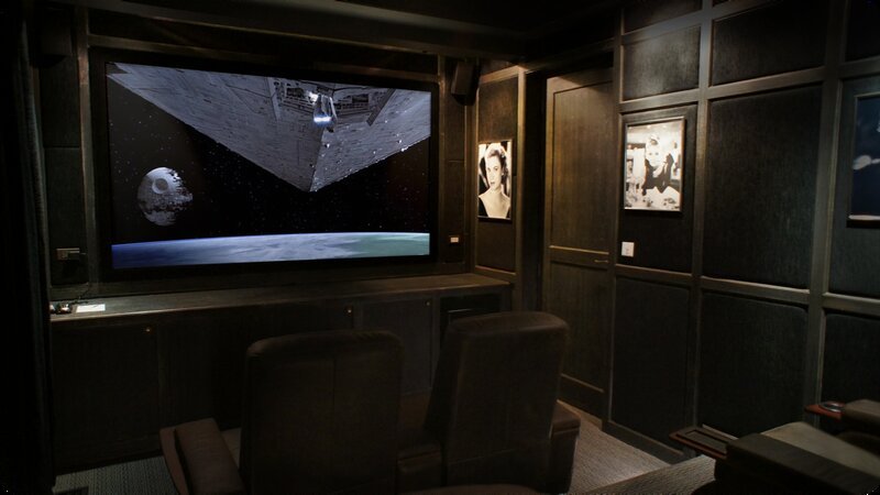 A home theater room with a large screen displaying a scene from space, including the Death Star and a spaceship. There are three black chairs facing the screen and framed black-and-white photographs on the dark wood-paneled walls.