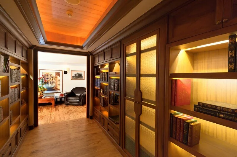 Wood-paneled library with built-in bookshelves, warm lighting, and a sitting area with in-ceiling speakers with a black armchair and a window with artwork outside.