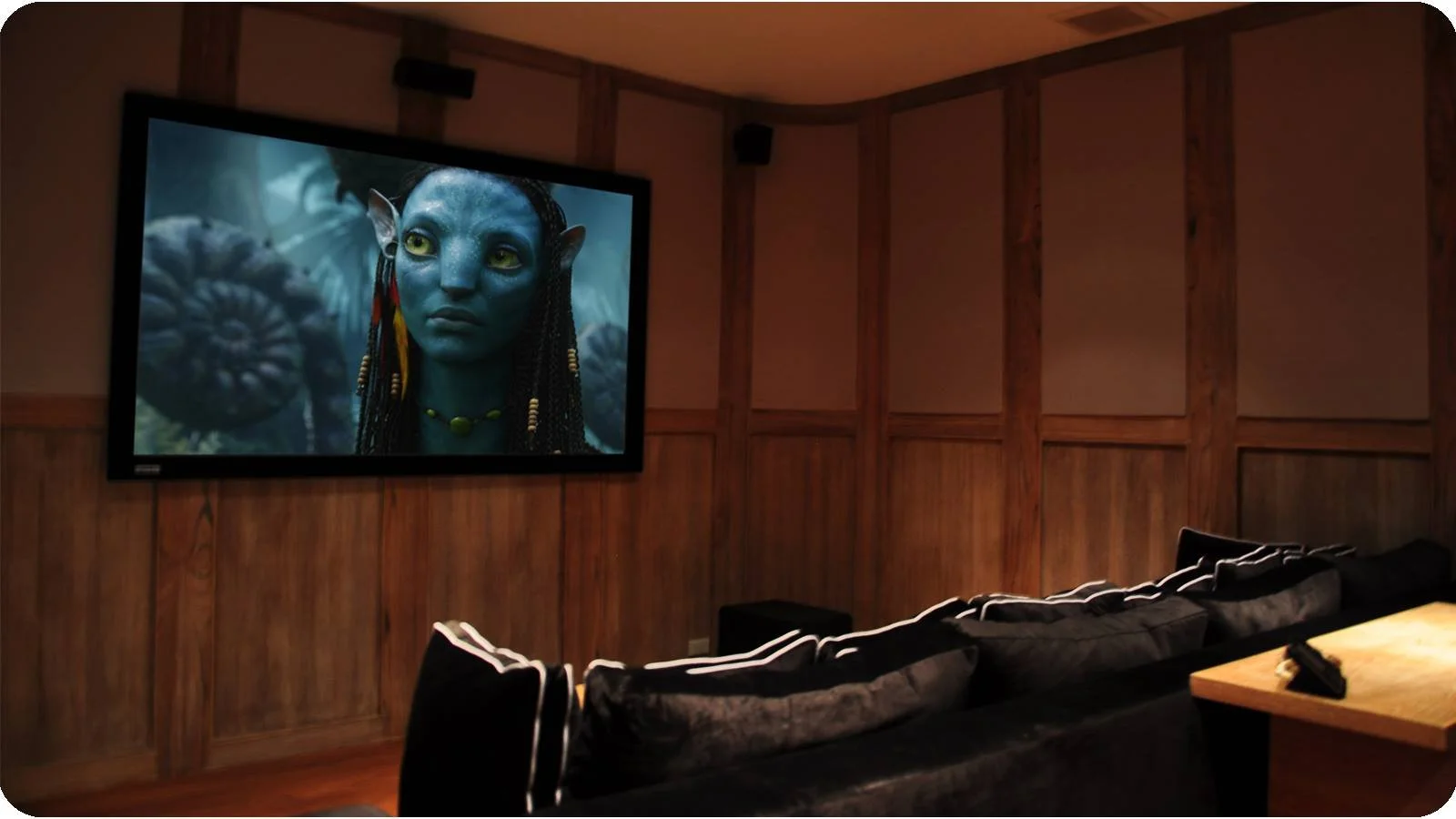 A movie theater room with a large screen displaying a scene from the film Avatar, featuring a blue alien character with braided hair and yellow eyes, in a wooden-paneled room with black sofas and a remote control on the table.