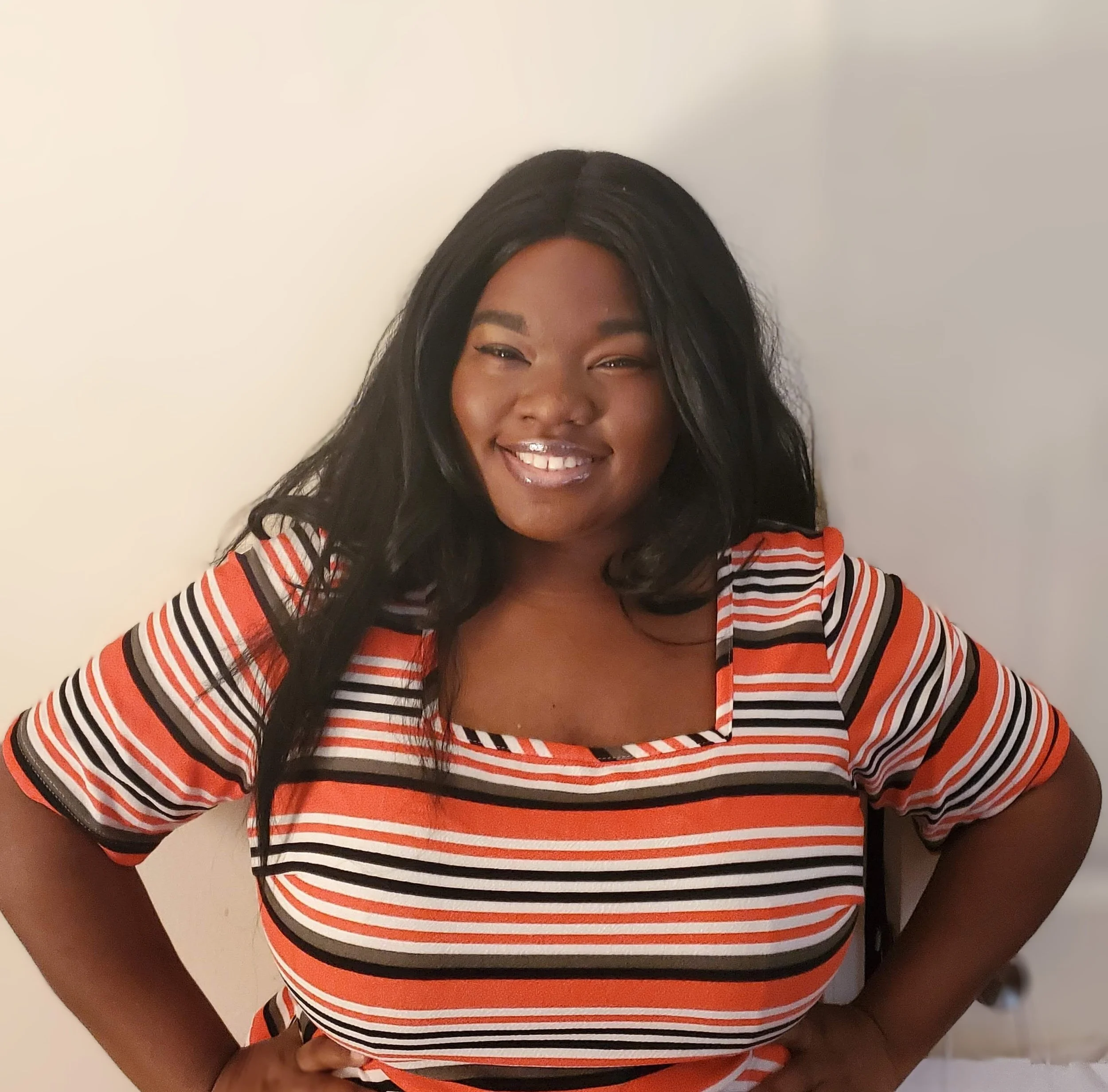 A woman with dark hair, smiling, wearing a striped orange, black, and white top, standing against a plain white wall.