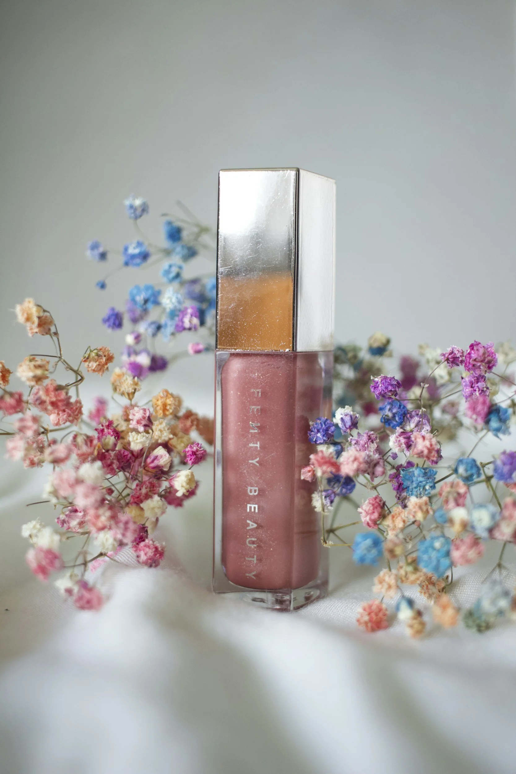 Pink lipstick in a clear tube labeled 'FREEDY BEAUTY,' surrounded by pink, purple, and blue small dried flowers.