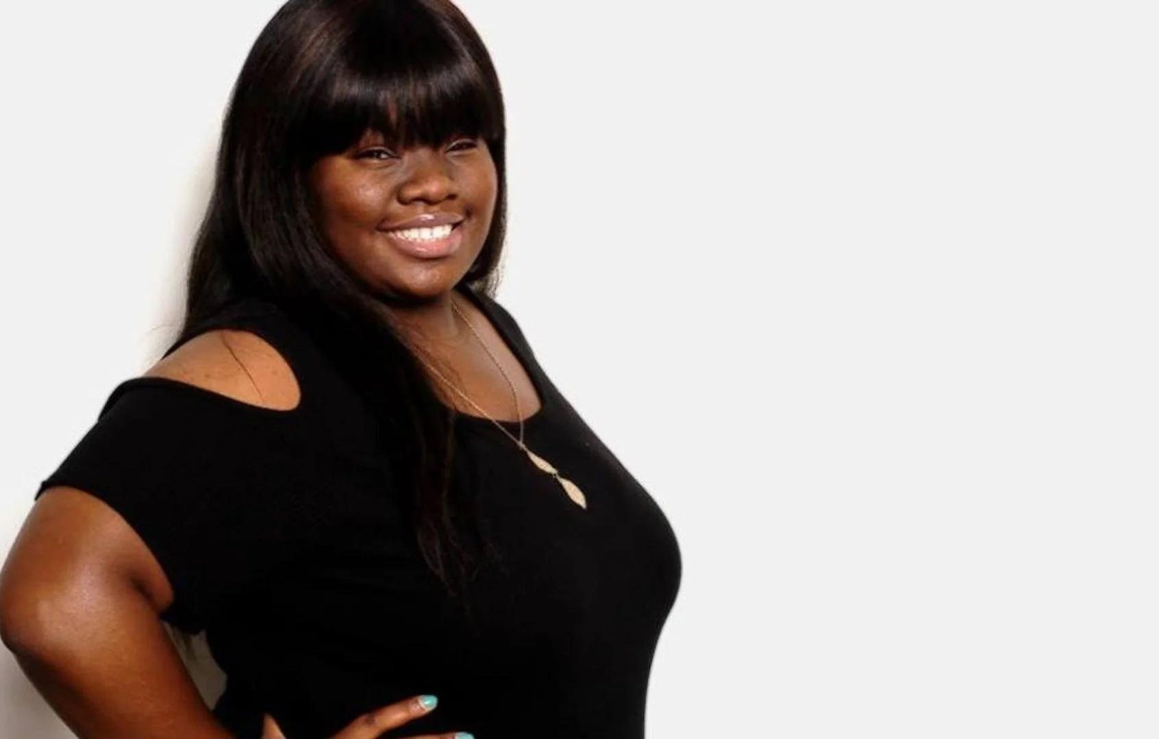 A smiling woman with dark hair wearing a black top with shoulder cutouts and seashell necklace, standing against a plain light background.