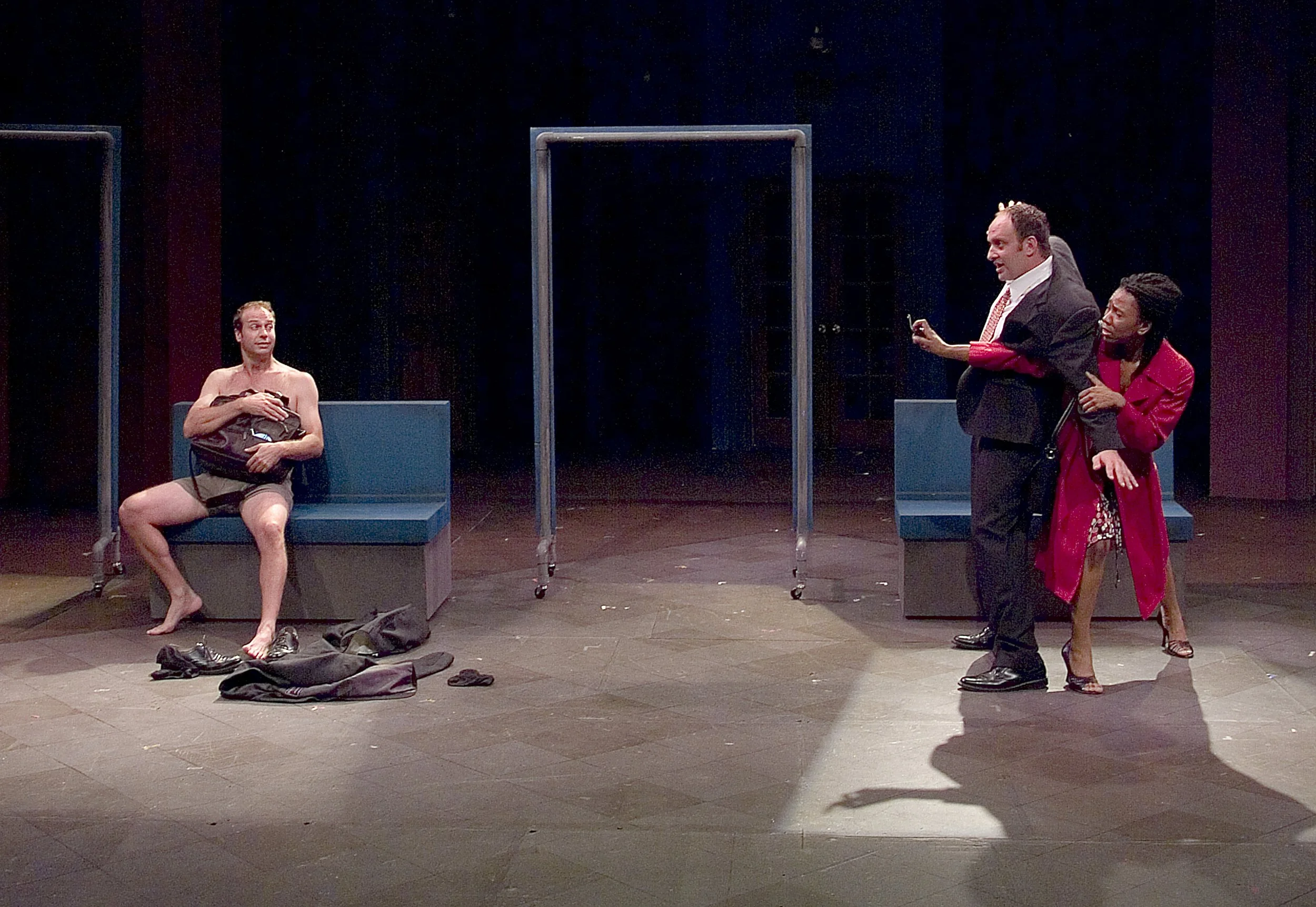 A theatrical scene with three actors on stage. One actor is sitting shirtless on a bench, holding his jacket, surrounded by scattered clothing and shoes. Two other actors stand to the right, one in formal attire and the other in a red coat, interacti
