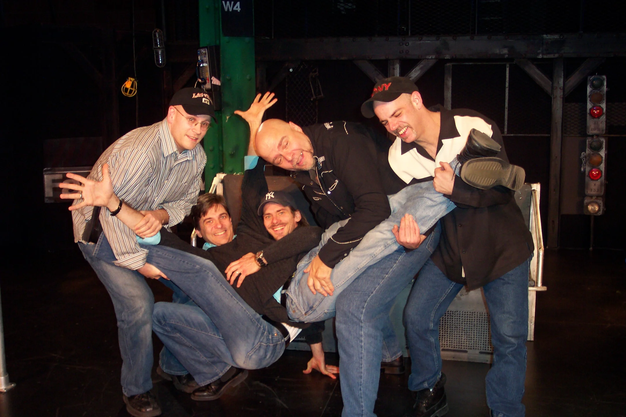 Five men playfully stacked on a bench, smiling and laughing, in a dark indoor setting with stage lights.