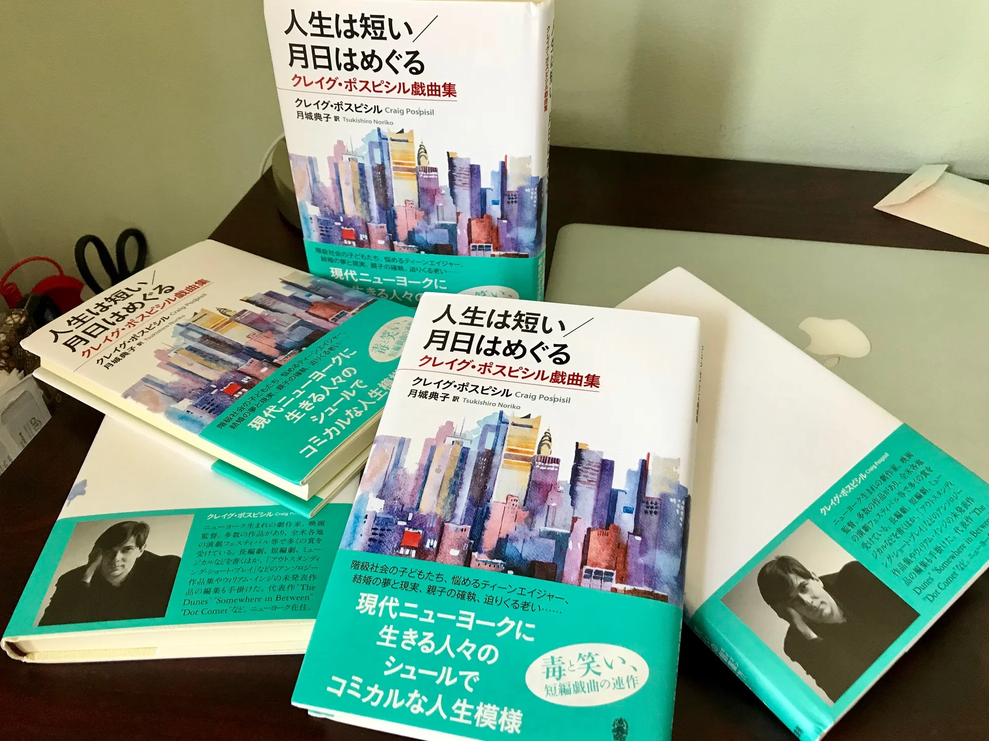Several copies of a Japanese book titled "人生は短い／月日はめぐる" by Craig Pospisil, with a watercolor illustration of a city skyline on the cover, placed on a dark wooden table.