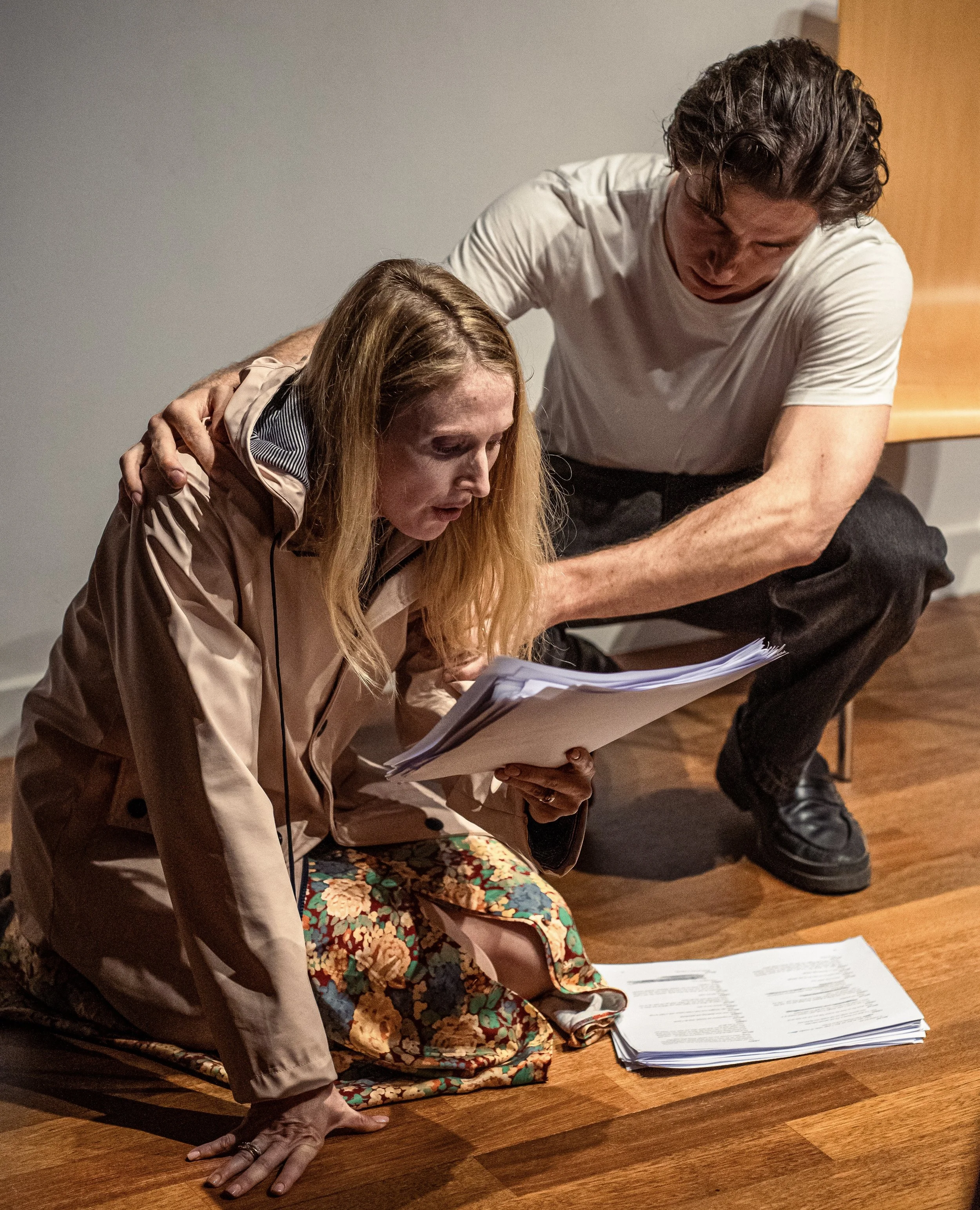 A woman is sitting on the floor holding papers, with a man leaning over her, appearing to check the documents.