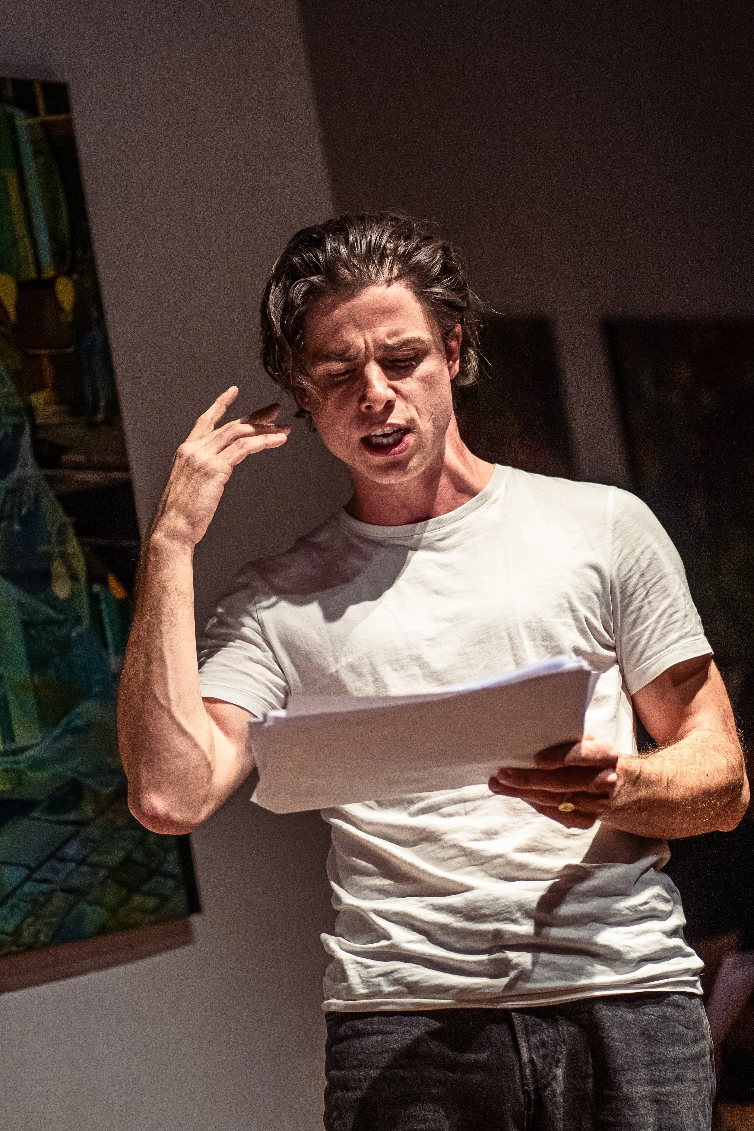 A young man with disheveled hair, wearing a white t-shirt, looks frustrated or angry as he reads from a stack of papers.