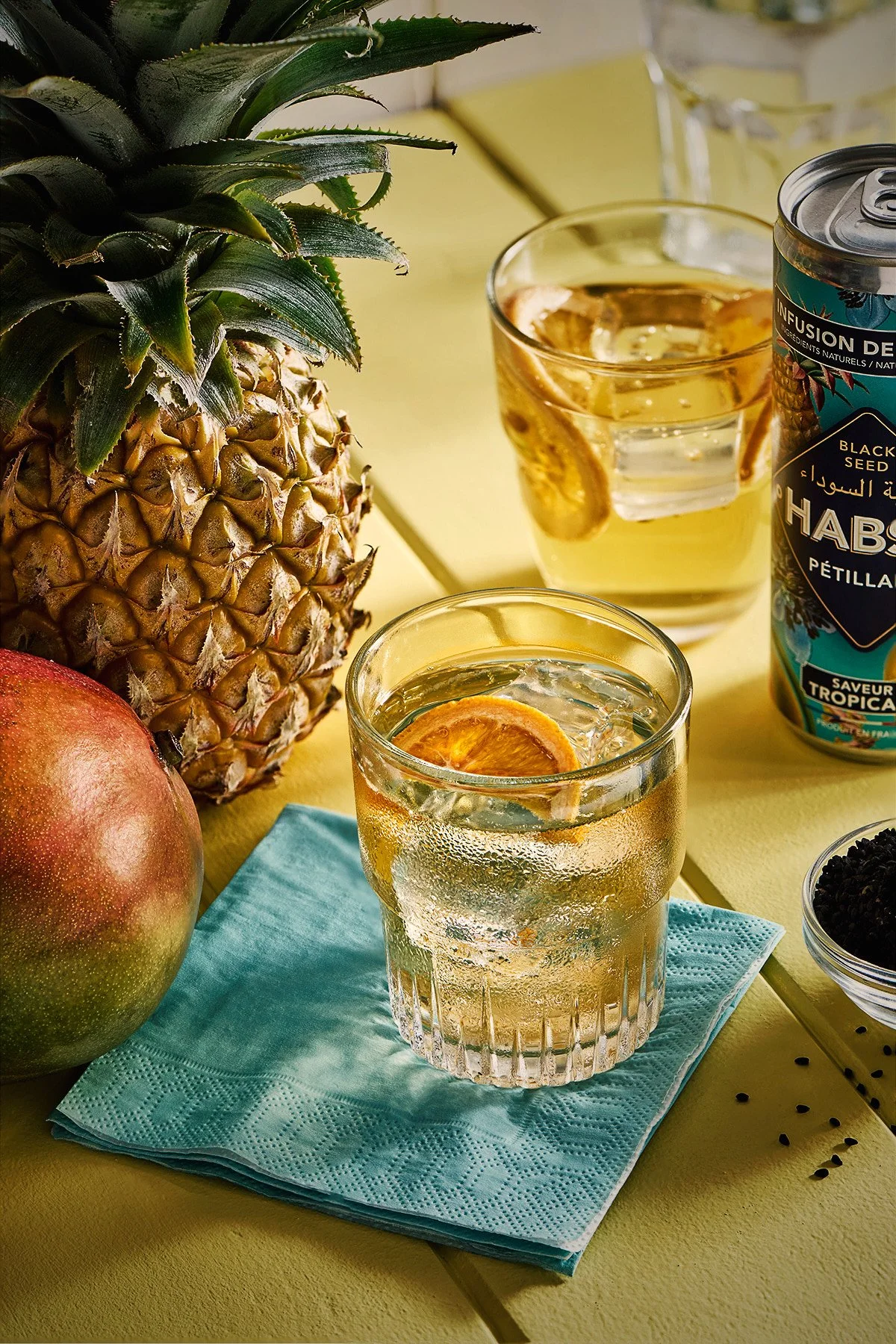 Une ananas, une mangue, et deux verres de boisson à base d'ananas avec des rondelles d'ananas, sur une nappe turquoise et un fond jaune.