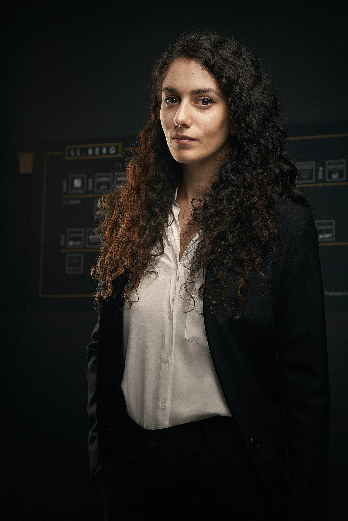 Femme avec cheveux bouclés, portant une veste noire et une chemise blanche, dans un environnement sombre avec un tableau de présentation en arrière-plan.