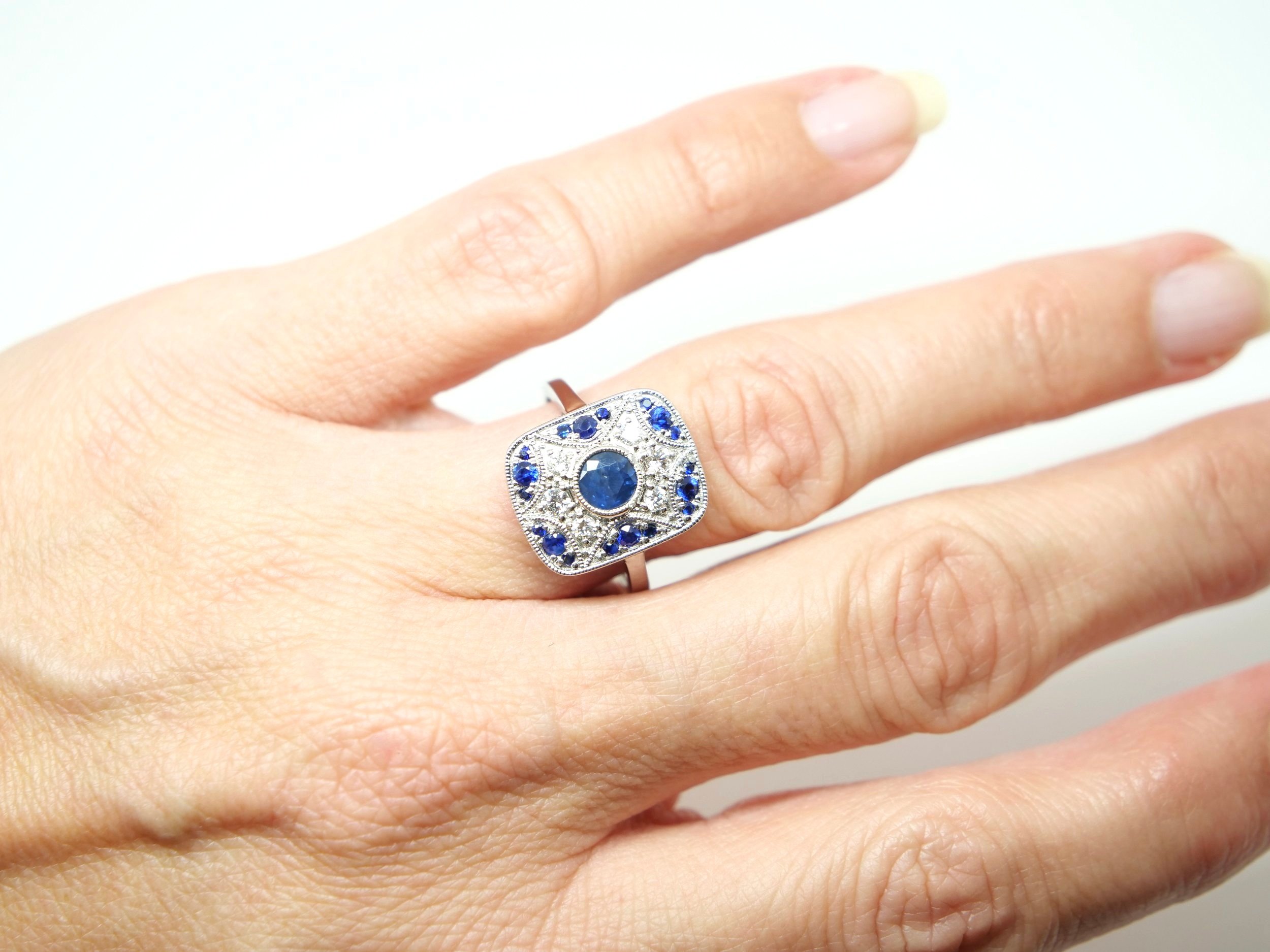 Unusual Art Deco style ring in 18ct gold, set with a central round cut sapphire, with further sapphires set into geometric design. Diamonds inset around central sapphire.