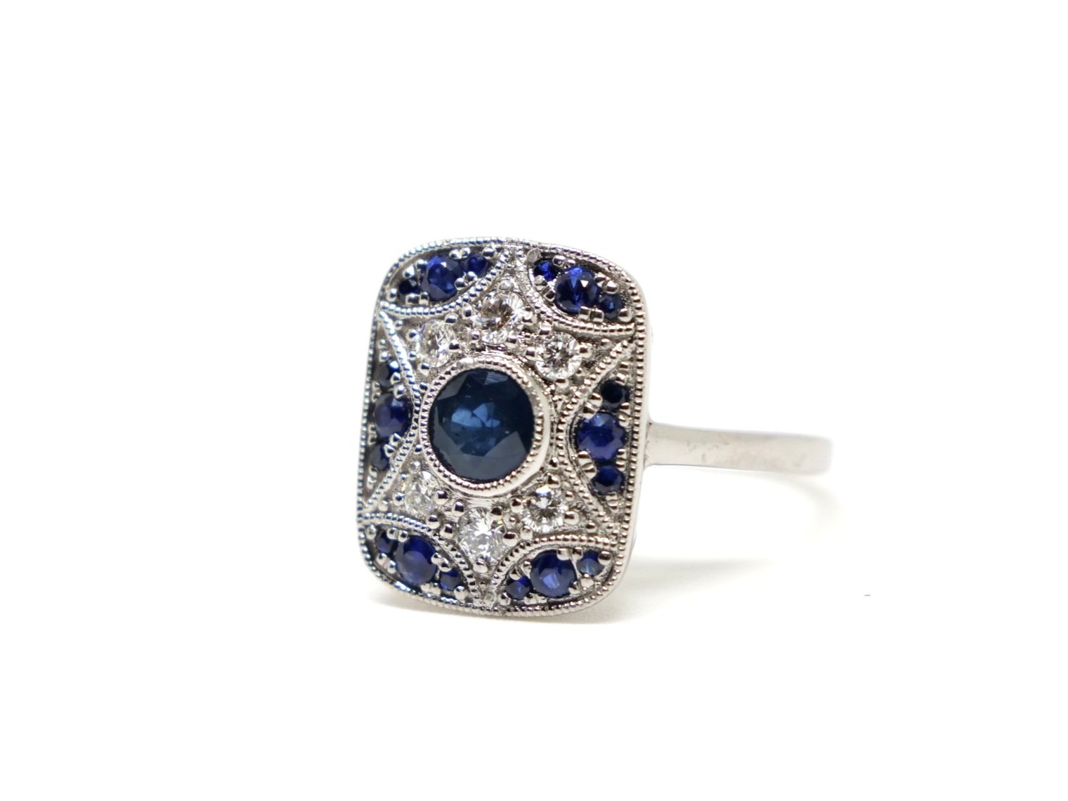 Unusual Art Deco style ring in 18ct gold, set with a central round cut sapphire, with further sapphires set into geometric design. Diamonds inset around central sapphire.